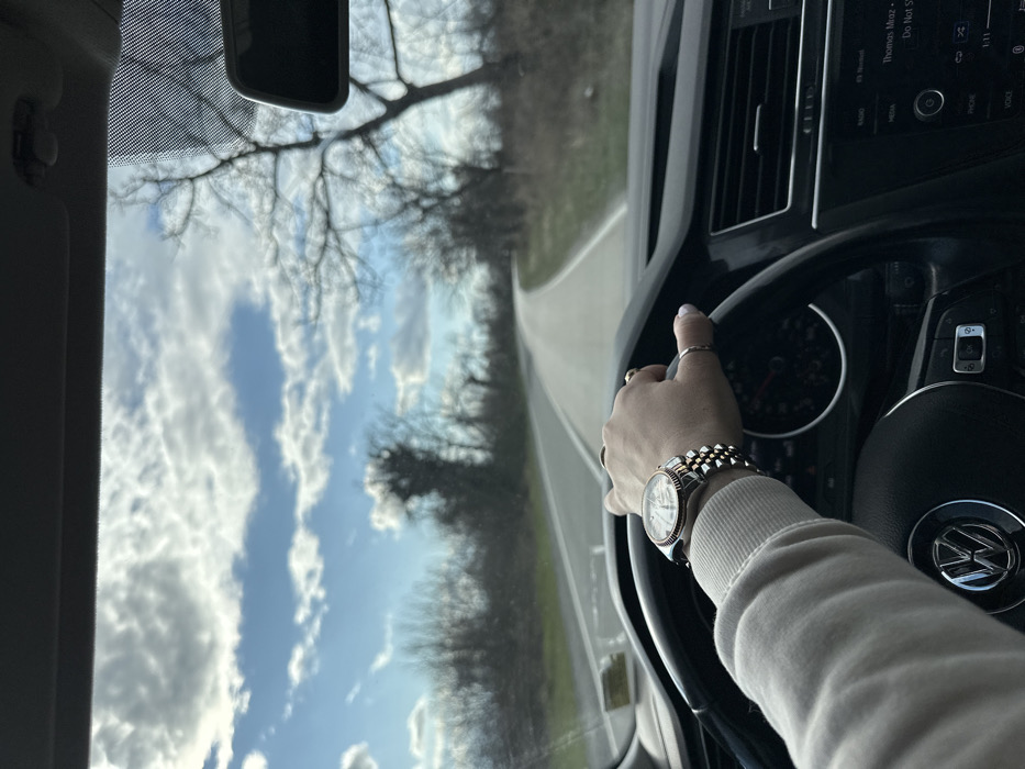 A first-person perspective from inside a vehicle in Kvareli, Georgia, shows a driver's left hand gripping a black multi-function Volkswagen steering wheel at the 9 o'clock position. The driver wears a light-colored, ribbed long-sleeved top. A two-tone metallic wristwatch with a fluted bezel is on the wrist, and several rings are visible on the fingers, which have light-colored nail polish. A portion of the vehicle's black dashboard, including air vents and a media display unit showing text such as "Thomas Mraz" and "Do Not S", is visible to the right of the steering wheel. Through the windshield, a two-lane asphalt road with white line markings extends forward, gently curving to the right in the distance. The road is bordered by dry grass and numerous bare deciduous trees, suggesting a late autumn or winter setting. The sky overhead is partly cloudy with patches of clear blue. A rearview mirror is partially visible in the upper right quadrant of the windshield.