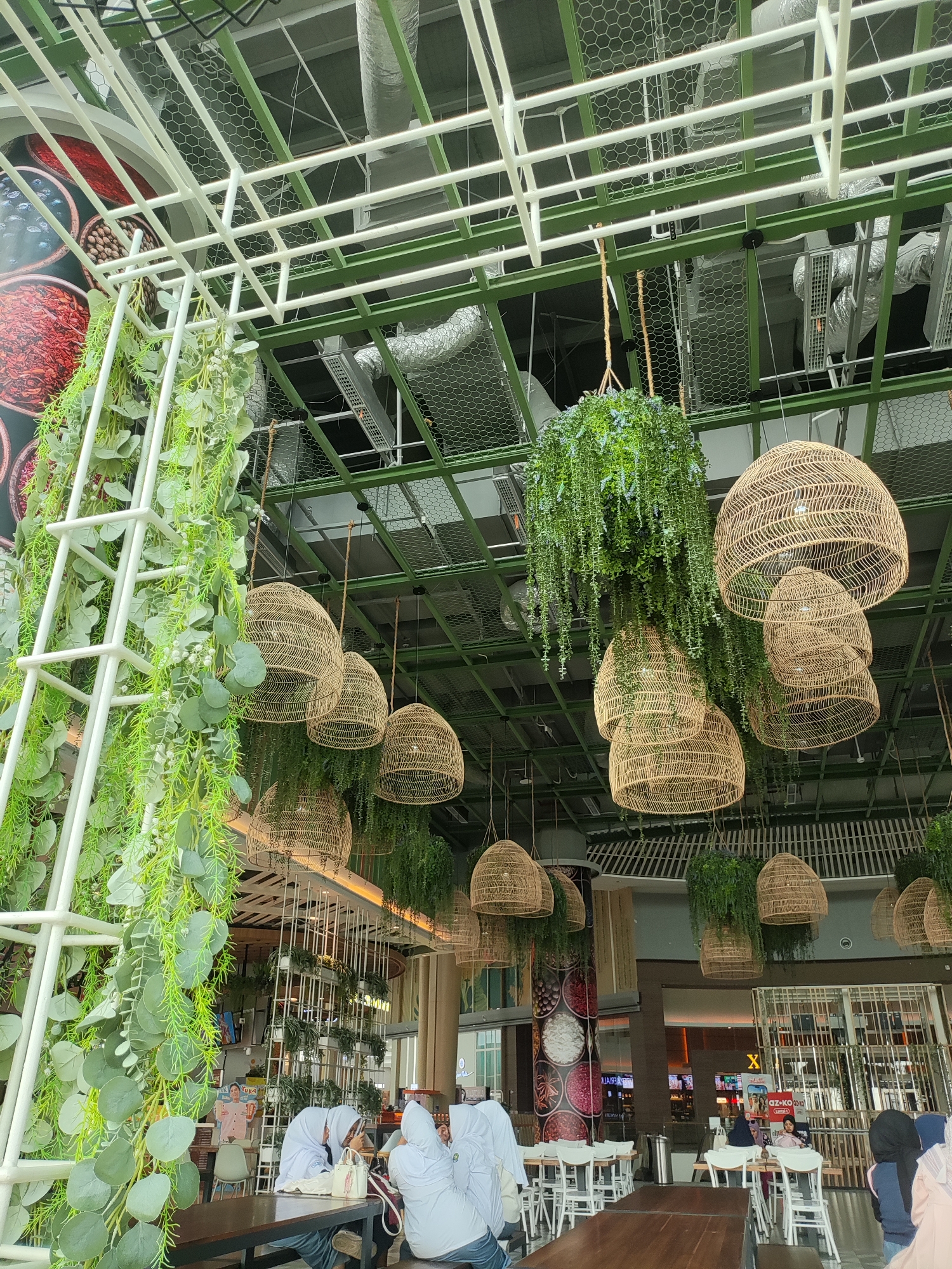 The image captures the interior of a modern, airy establishment, likely a restaurant or cafe, with a high ceiling featuring exposed ductwork and green metal beams. Several woven pendant lamps, shaped like inverted baskets, hang from the ceiling, interspersed with lush hanging plants, creating a natural, bohemian ambiance. A prominent white lattice structure on the left is adorned with cascading artificial greenery, adding to the botanical theme. In the background, tables and chairs are set up, with several patrons seated, including a group of individuals wearing white hijabs and tops, suggesting a cultural context. The overall impression is one of a stylish, contemporary space that blends industrial elements with organic decor. The reference to Lambangjaya, Indonesia, indicates the geographical setting of this establishment.