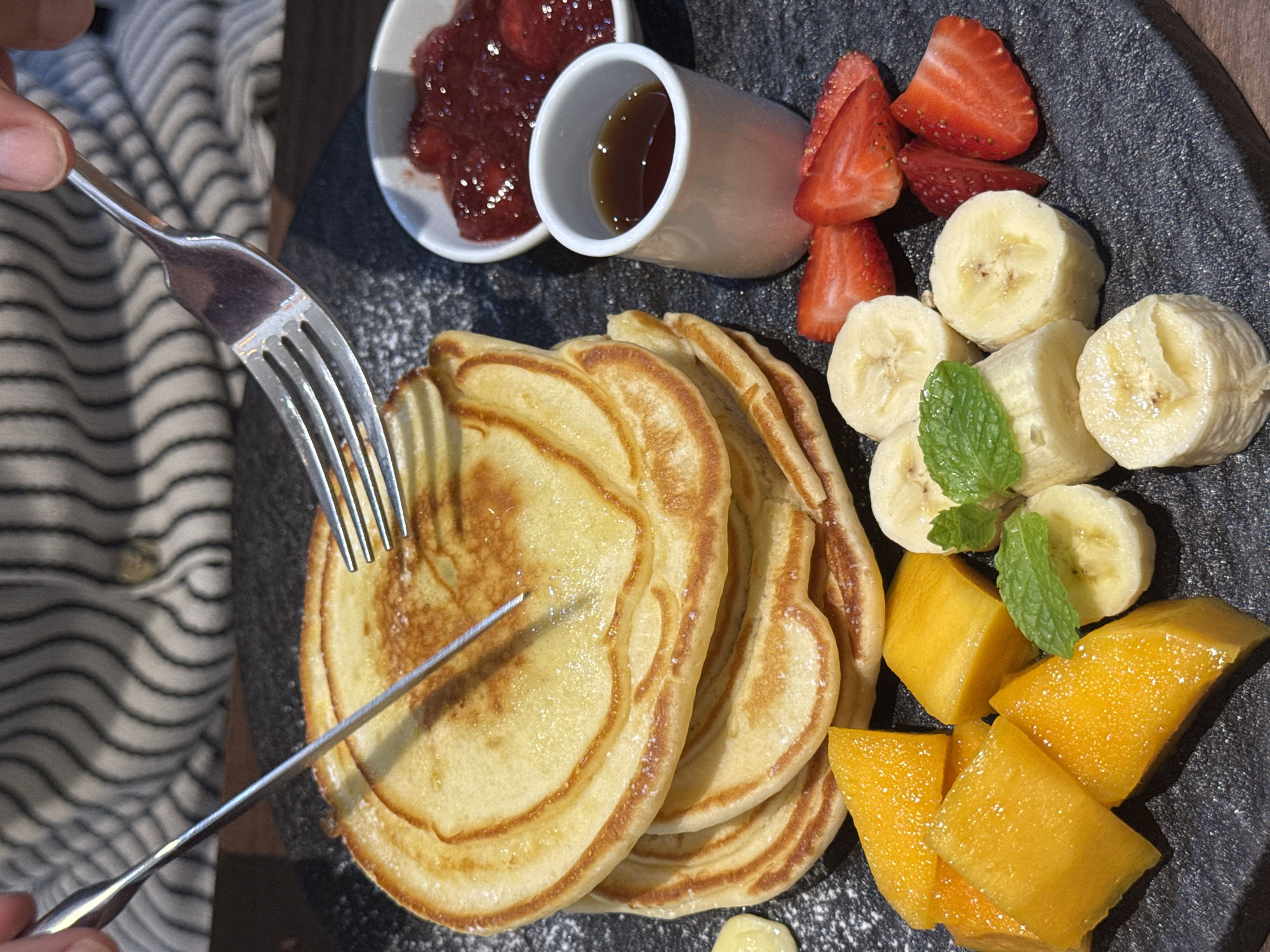 The image shows a plate of food, including pancakes, sliced strawberries, sliced bananas, and diced mango. There are also two small containers, one with a red jam-like substance and the other with a dark liquid, likely syrup. A fork and knife are being used to cut the pancakes. The meal is presented on a textured dark gray plate. The background includes a striped fabric or clothing. While the location in Đà Nẵng, Vietnam is requested, there is no direct indication of the city present in the image.