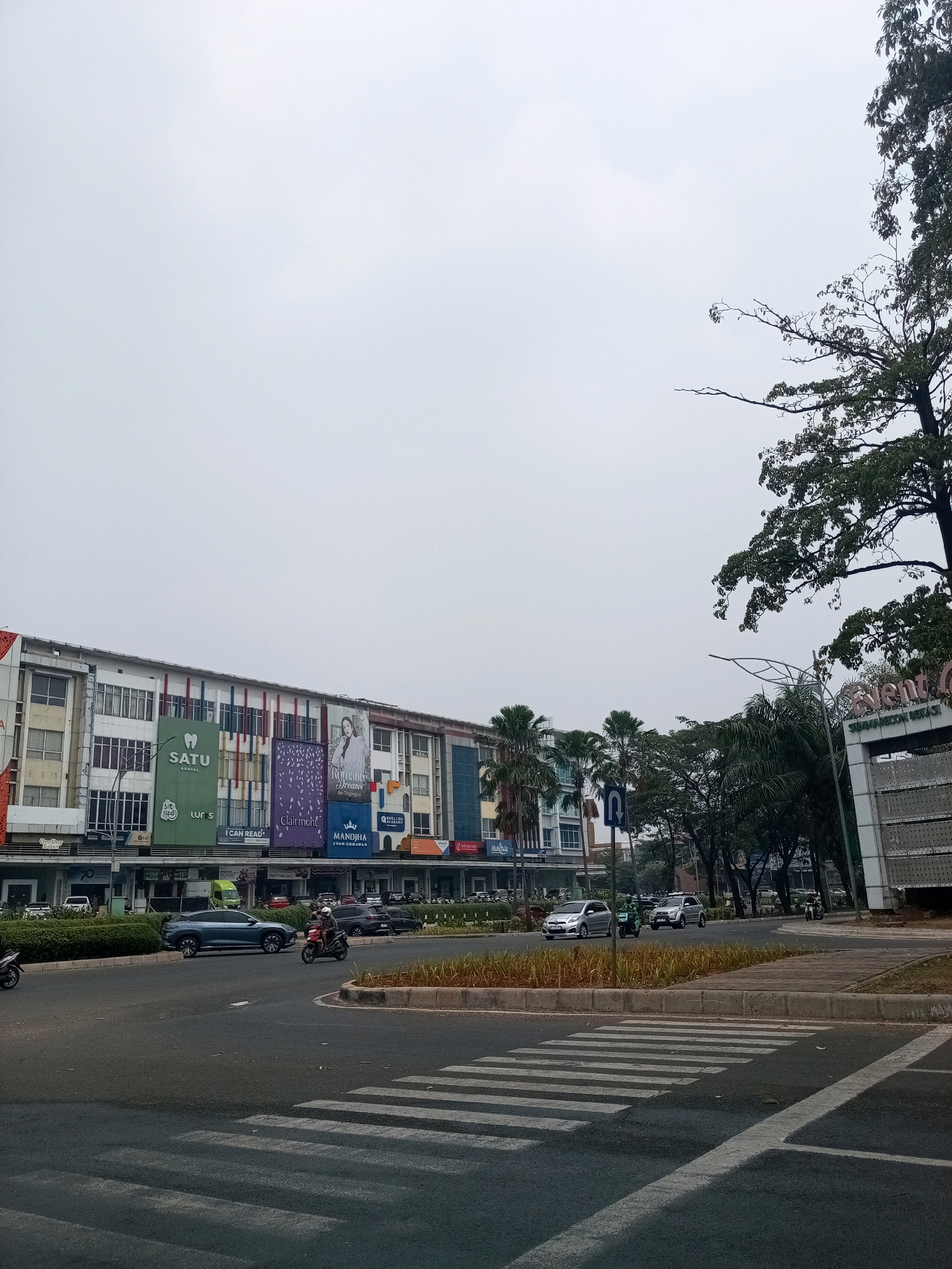 In Bekasi, Indonesia, a street scene unfolds under an overcast sky. The foreground features a pedestrian crossing on a paved road. To the left, a row of multi-story buildings displays various storefronts and advertisements, including "SATU DENTAL," "LUR'S," and "Clairmont." A dark-colored SUV is stopped on the road, with a person on a motorcycle approaching it. Further down the street, more vehicles are visible. Palm trees and other foliage line the side of the road. To the right, a modern building with the text "Event" is partially visible. The scene captures a typical urban environment with daily activities.
