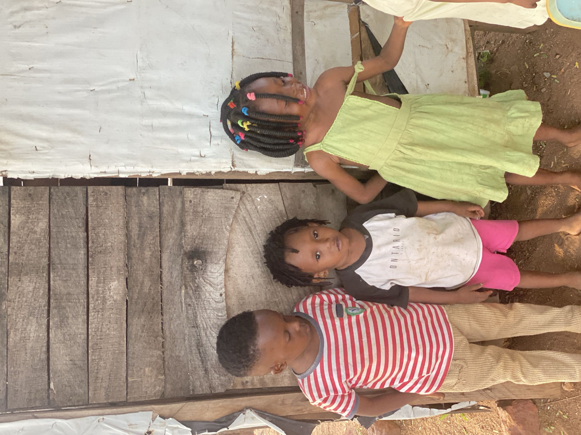 Three children are positioned on a wooden surface, likely a porch or platform, adjacent to a wall constructed of wood and white sheeting. The children are lying horizontally in the frame. The first child is wearing a red and white striped shirt and light tan pants. The second child is wearing a white shirt with "ONTARIO" printed on it, with black sleeves, and pink shorts. The third child, situated at the top of the frame, is wearing a light green dress with braided hair adorned with colorful hair accessories. The environment consists of soil and partial structures, implying an outdoor setting. The location is indicated as Adankolo, Nigeria.