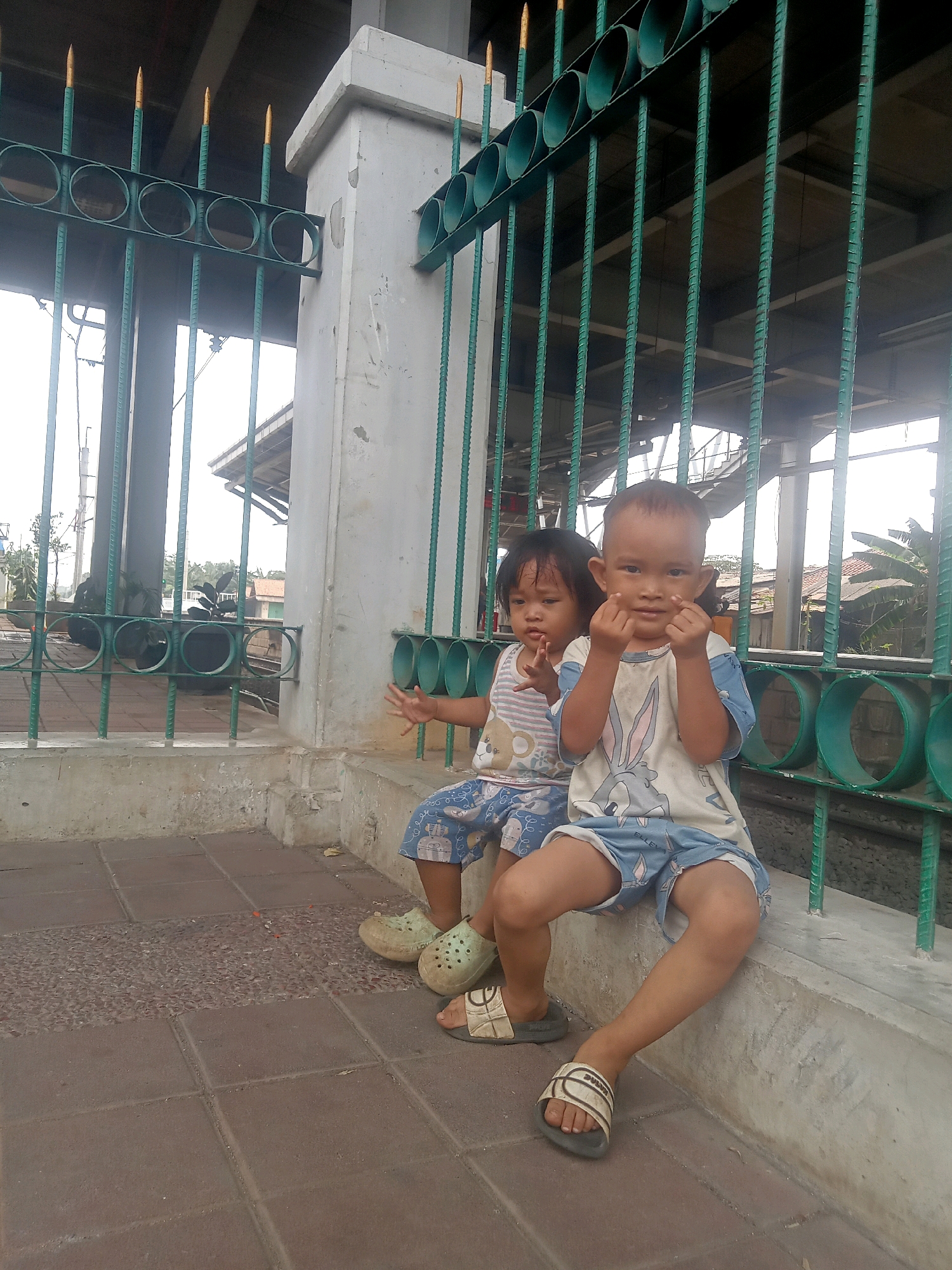 Two young children, a boy and a girl, are sitting on a concrete ledge outdoors. The boy, who is on the right, is wearing a light-colored t-shirt with a cartoon bunny graphic and blue shorts with a white pattern. He is looking at the camera with his hands held up near his face, making a gesture. The girl, on the left, is wearing a striped t-shirt with a bear graphic and blue shorts with a similar white pattern. She is looking slightly off to the side and has one hand raised. They are both wearing sandals. Behind them is a green metal fence with circular openings and pointed tops, attached to a concrete pillar. The ground in front of them is paved with square tiles and a section of textured material. In the background, there are buildings and vegetation, suggesting an urban or suburban environment.
