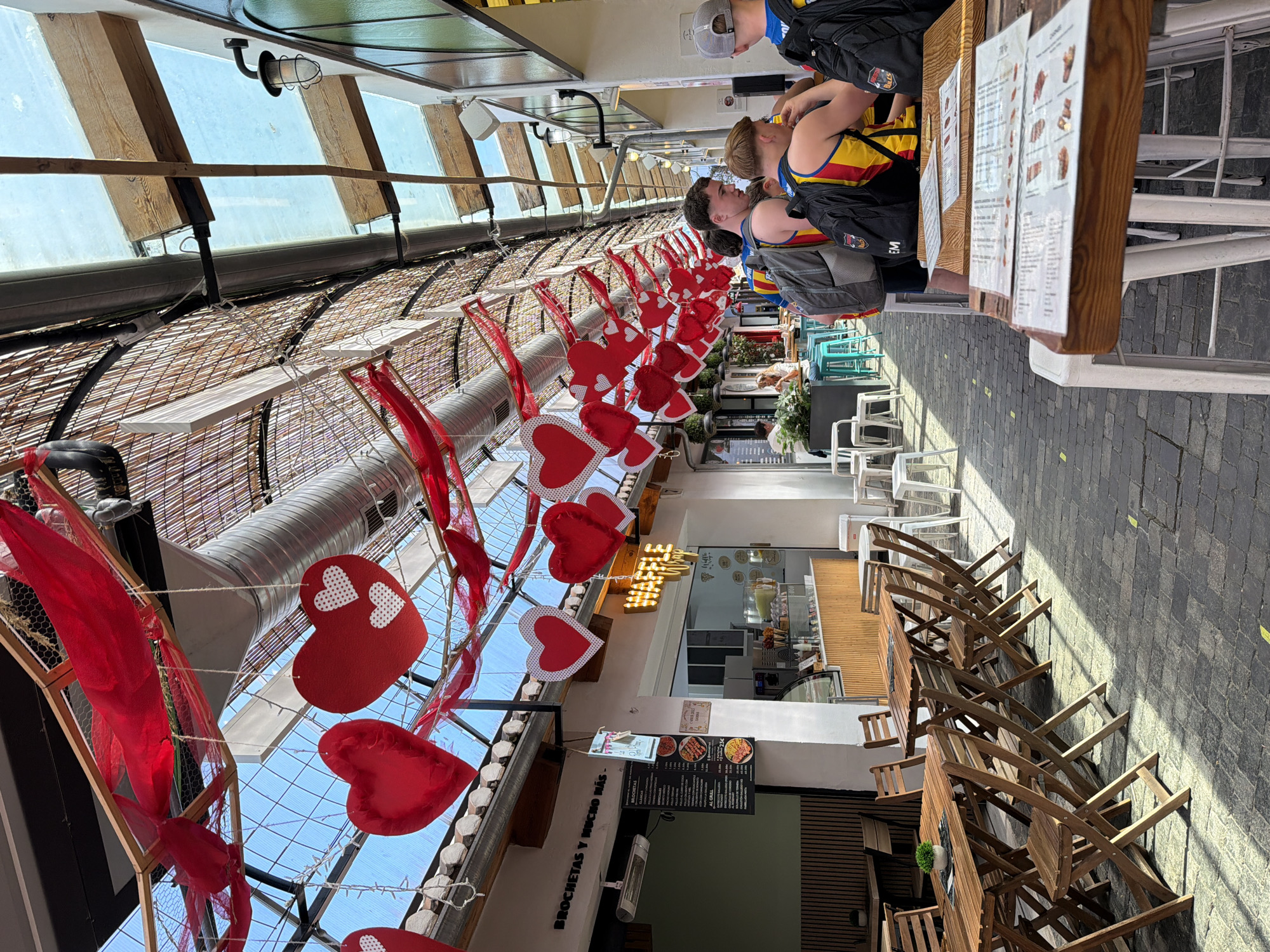 The image depicts an outdoor seating area of a restaurant, possibly in Dénia, Spain, given the presence of a menu sign with the word "BROCHETAS," a Spanish term for skewers. The seating area is decorated with strings of red heart-shaped ornaments, possibly for Valentine's Day or another romantic occasion. Above, a partially covered roof provides shade, with a view of the sky visible through the openings.
Several patrons are seated at tables. The menu is visible on the tables. Seating consists of a mix of white chairs and wooden folding chairs. The floor is paved with grey brick. The scene appears to be a sunny day, as evidenced by the shadows cast on the pavement.