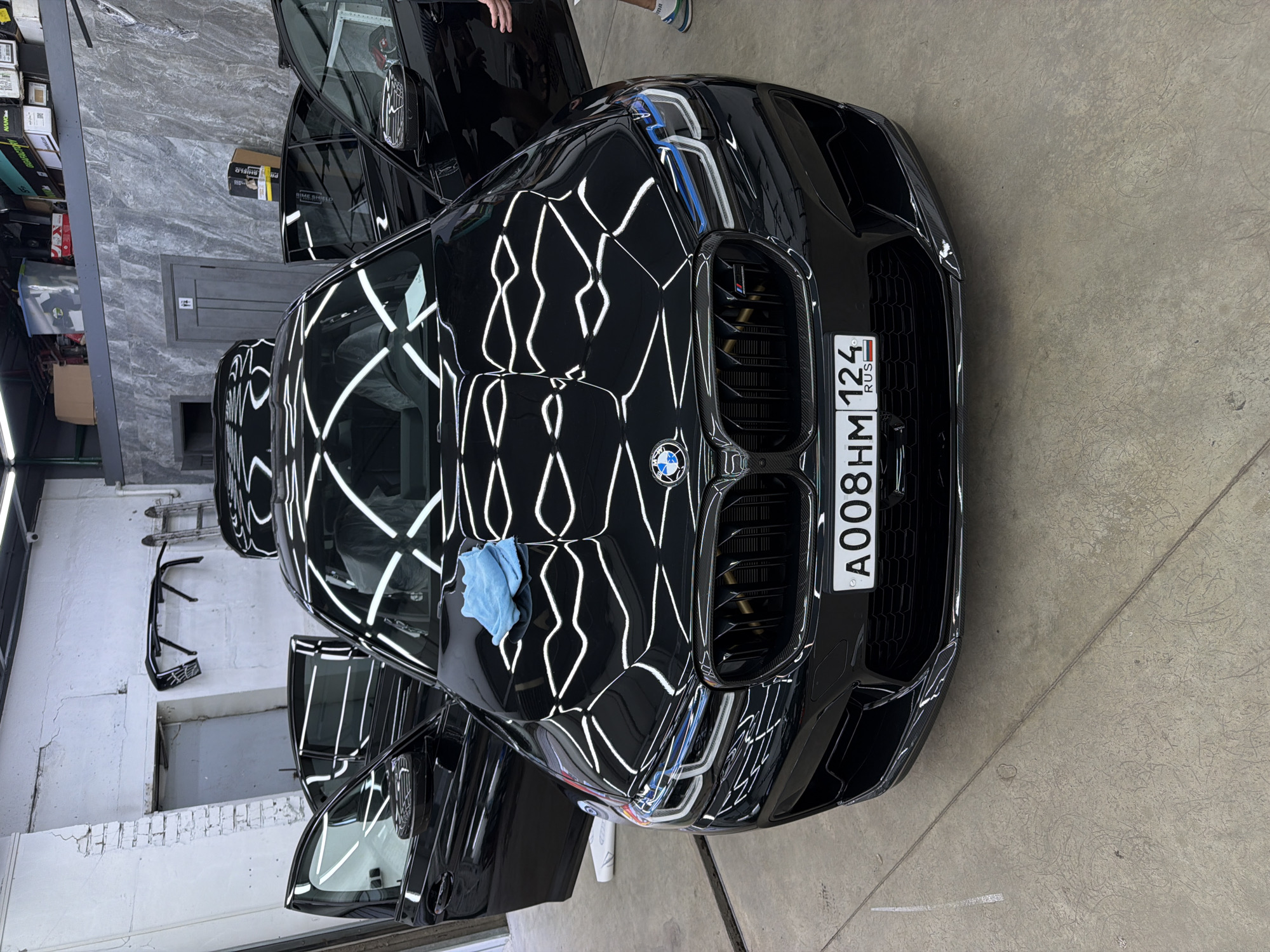 A black BMW M5, its doors open, is parked inside a commercial garage, likely a detailing or repair shop. The car's hood is covered with a light blue microfiber cloth, and the interior is wrapped in plastic, suggesting recent detailing or preparation for storage. The garage floor is concrete, marked with white lines. In the background, shelves are stacked with boxes, and a ladder leans against a white brick wall. A person, partially visible on the right, is standing near the car. The license plate of the car, visible in the foreground, reads "A008HM 124 RUS". This image was captured in Krasnoyarsk, Russia.