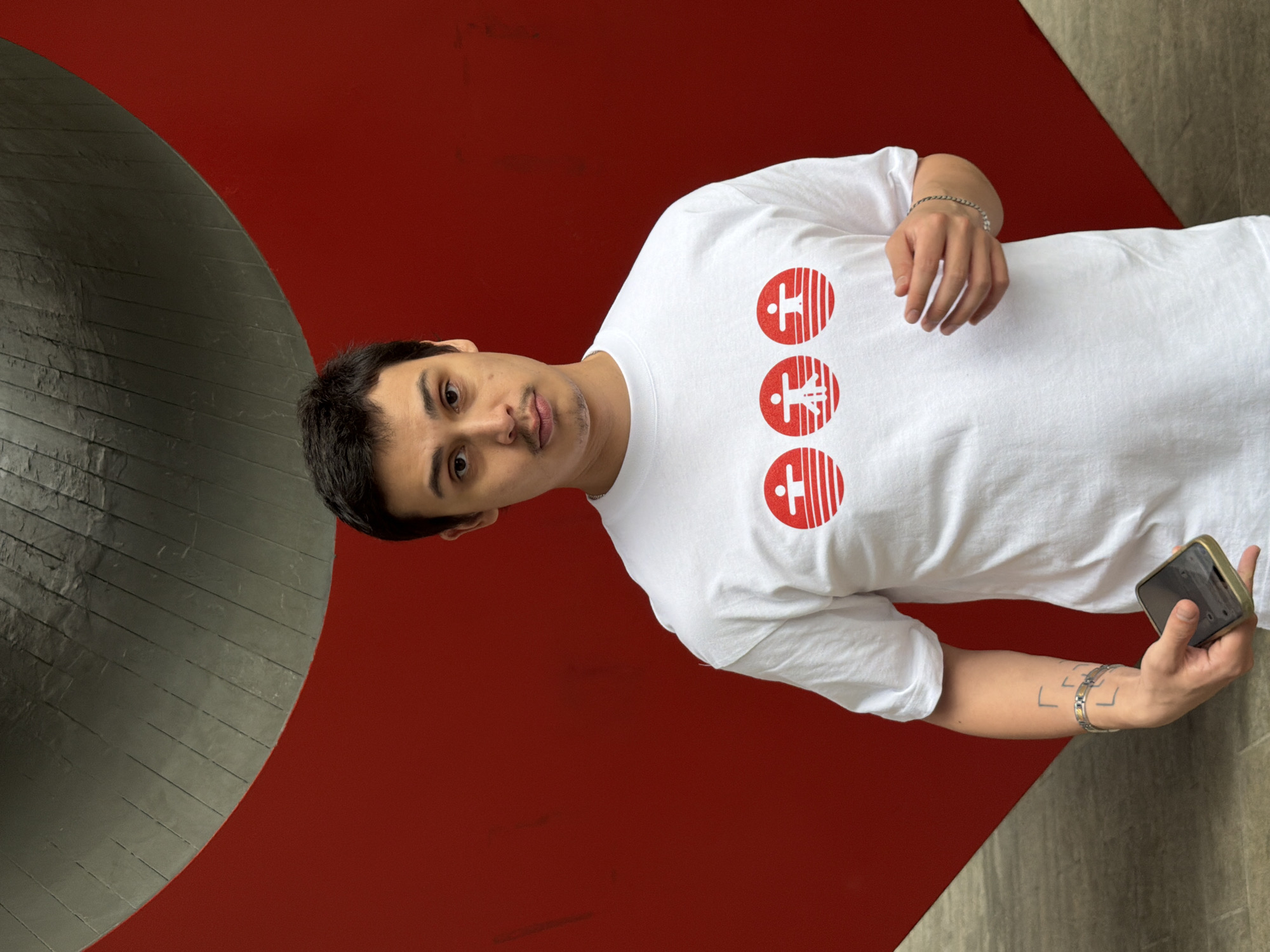 A man stands in front of a large sculpture or architectural detail that is painted in two colors, gray and red. The man is wearing a white t-shirt with three red circles printed on it. Each circle contains a different icon. He is also wearing bracelets on both wrists. He is holding a cell phone in his right hand. The man has dark hair and a light complexion. The scene appears to be outdoors. The subject is oriented so that the right side of the frame is pointing down.
While the exact location cannot be determined from the image alone, the presence of the large sculpture could indicate a location within New York City, United States of America, which is well known for its public art installations and unique architectural features.