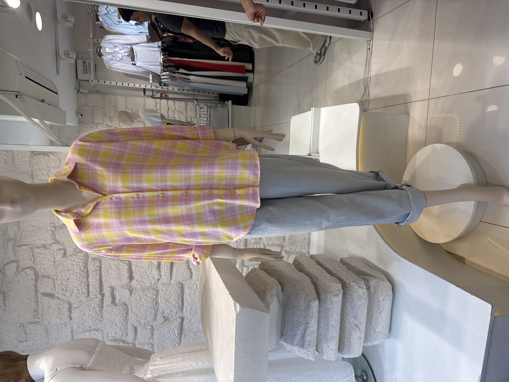 A mannequin wearing a pink and yellow plaid button-down shirt and light blue cuffed jeans is positioned in a store. The mannequin is standing behind a white, textured display table. In the background, a rack of clothing and a mirror are visible. A man wearing a black shirt and khaki pants is reflected in the mirror. The scene appears to be a clothing store in Gungoren, Turkey.