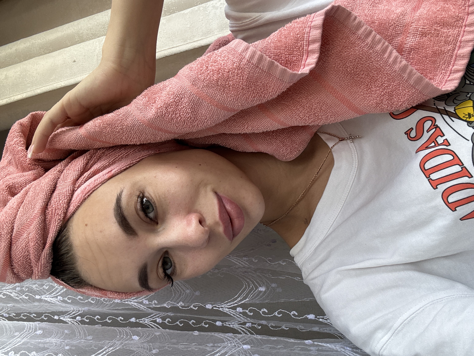 This media file, captured in Chaikovsky, Russia, features a young woman looking directly at the camera while lying down. Her head is wrapped in a pink terry cloth towel, suggesting she has recently washed her hair. A hand is visible at the top of the frame, holding the towel in place.
The woman has a calm and slightly smiling expression, with her eyes open and clearly visible. She has well-defined eyebrows and prominent eyelashes. She is wearing a white short-sleeved t-shirt, on which part of the red text "ADIDAS" is visible, along with what appears to be a graphic or cartoon character to its right. A thin gold chain with a small, sparkling cross pendant rests on her collarbone.
She is positioned against a sheer white fabric with intricate white embroidered patterns, possibly a curtain or bedspread. The background shows a light-colored, possibly textured surface, which could be a wall or steps, suggesting an indoor environment. The lighting indicates it is daytime. The overall scene conveys a relaxed and casual moment in a domestic setting.