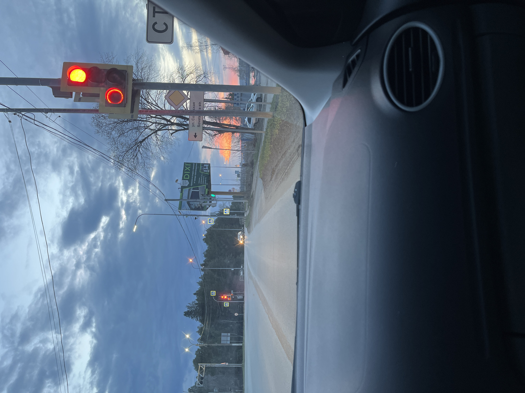 The image presents a driver's perspective from inside a car stopped at an intersection in Chaikovsky, Russia. The scene depicts either an early morning or late evening hour, characterized by the illuminated streetlights and a vibrant sunset or sunrise painting the horizon with orange and pink hues, contrasting with the dark, cloudy sky overhead.
The car is halted by a red traffic light, which is prominently visible on the left, accompanied by a smaller illuminated red circular signal below it. Several road signs are apparent, including one pointing left with "ул. Советская" (Sovetskaya str.) and its English translation "Sovetskaya str.", as well as a large green sign for the "DIXI" supermarket. Yellow diamond-shaped warning signs are also scattered around the intersection.
The street ahead, partially lit by streetlights, shows clear road markings. Bare trees line the roadside, suggesting a colder season. Overhead power lines crisscross the upper part of the frame. A portion of the car's dashboard and an air vent are visible on the right, framing the view.
Visible text includes "CT" (likely part of a larger sign), "A" on the warning signs, "DIXI", "ул. Советская", "Sovetskaya str.", and fragments of text on the DIXI sign such as "строения из сэндвич-п" (buildings from sandwich panels), "от завод" (from factory), and "одител" (driver).