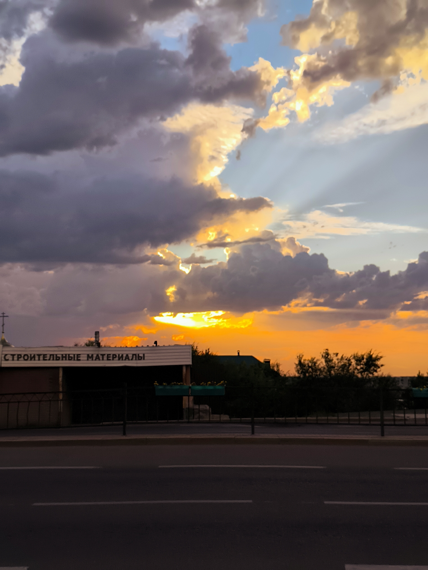 A dramatic sunset over a street in Qarağandı, Kazakhstan, is captured in this vertical, medium shot. The sky is ablaze with vibrant orange and yellow hues where the sun breaks through dark, billowing clouds, casting ethereal light rays. In the foreground, a paved road with white lane markings runs horizontally, with a sidewalk and a dark fence lining the street. Behind the fence, a building with the Cyrillic text "СТРОИТЕЛЬНЫЕ МАТЕРИАЛЫ" (Building Materials) is visible, topped by a small dome with a cross. Silhouetted trees and structures are scattered across the background, adding depth to the scene. The overall impression is one of natural beauty and urban life coexisting.