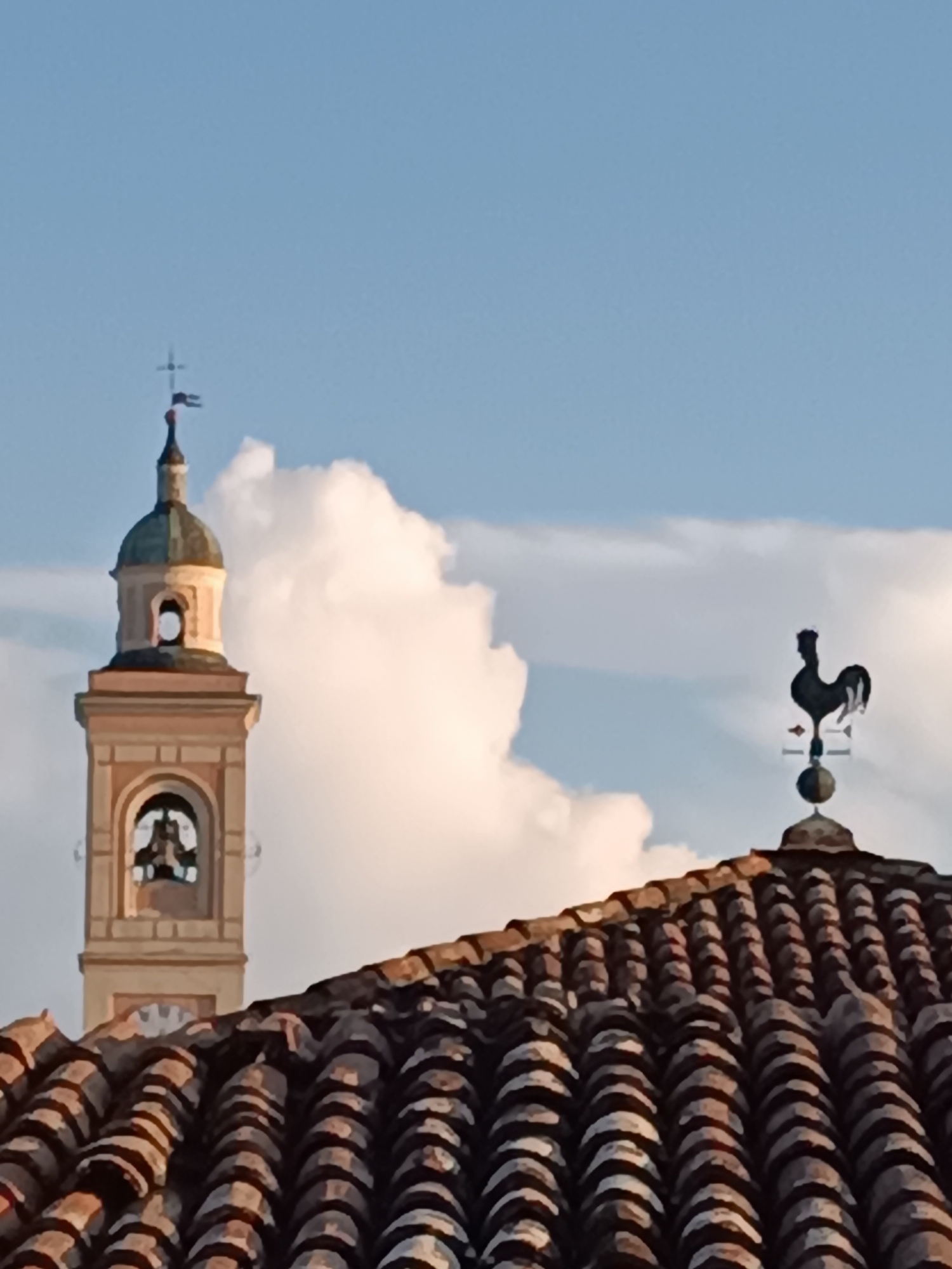The image shows a close-up view of a bell tower and a weather vane on a tiled roof, against a sky with clouds. The bell tower, likely part of a church in Viadana, Italy, is light brown with a green dome and features an open archway with a bell inside. A cross tops the tower. To the right, a black weather vane shaped like a rooster stands atop a dome. The foreground is dominated by the textured surface of a brown tiled roof. The scene is illuminated by sunlight, casting shadows and highlighting the architectural details. No people are visible in the image.