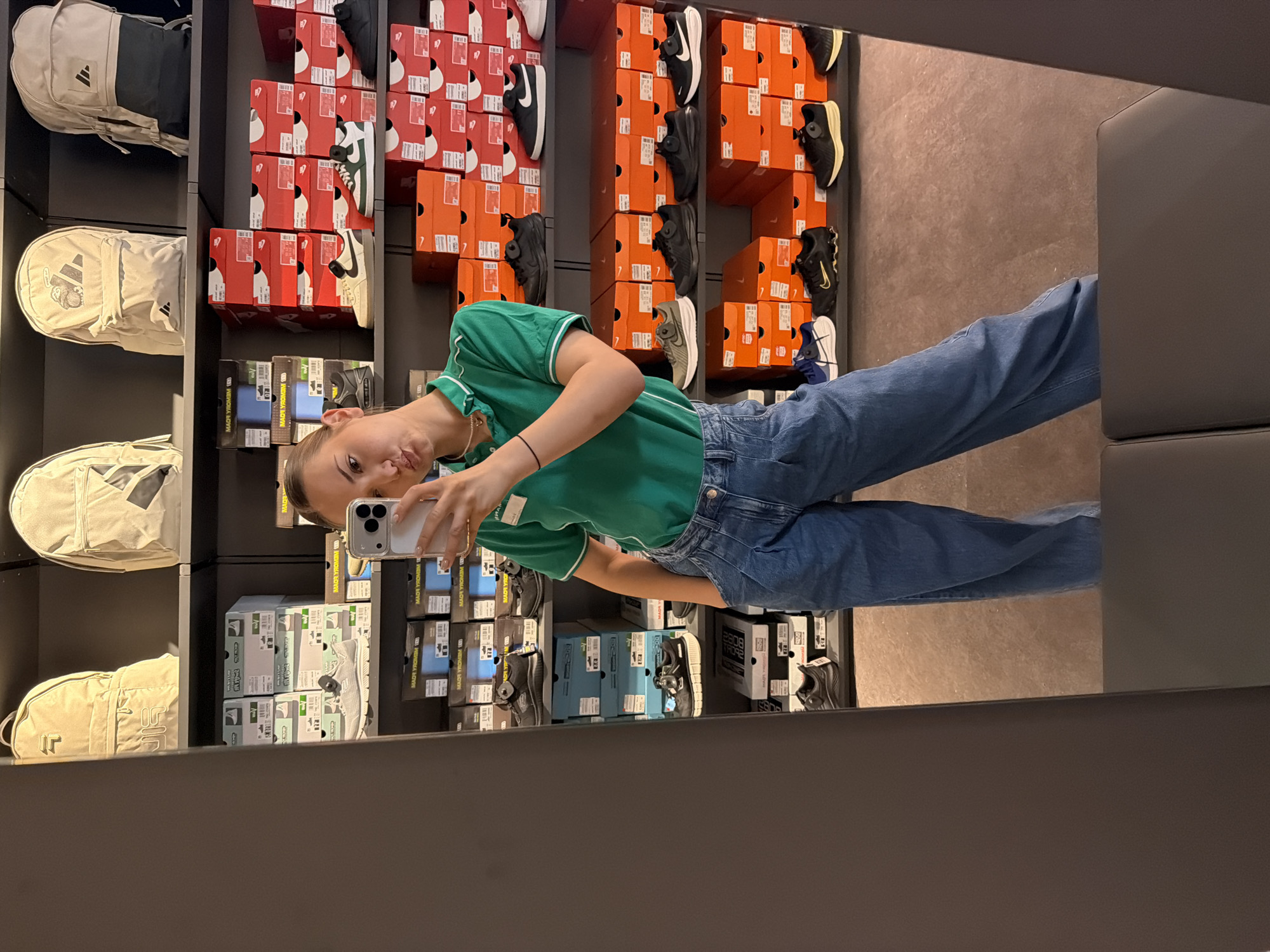 A young woman is taking a selfie in a mirror inside a shoe store. She is wearing a green polo shirt and blue jeans. The store has shelves of shoes and shoe boxes on the walls. The text "MAOR YROMIM" is visible on some of the boxes. The location is likely a shoe store, potentially in Sofia, Bulgaria.