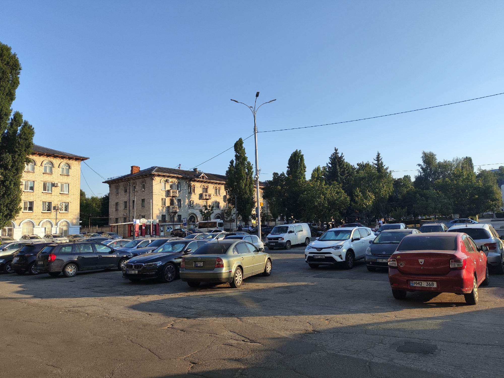 A wide-angle, eye-level outdoor shot captures a parking lot filled with various cars under a clear blue sky. In the background, two multi-story buildings with light-colored facades and dark roofs are visible, suggesting an urban setting. Trees line the street and border the parking area. A lamppost with three lights stands tall in the middle ground, with electrical wires spanning across the frame. The parking lot itself is paved with asphalt, showing signs of wear and tear. Several cars are parked in rows, including a red Dacia Logan with the license plate MHQ 368, a light green Volkswagen Passat, a white Toyota RAV4, and a dark gray Dacia Logan station wagon. People are present in the background, some walking near the buildings. The overall impression is a typical city scene on a bright, sunny day. This scene is likely in Chișinău, Moldova, given the presence of Moldovan license plates and the architectural style of the buildings.