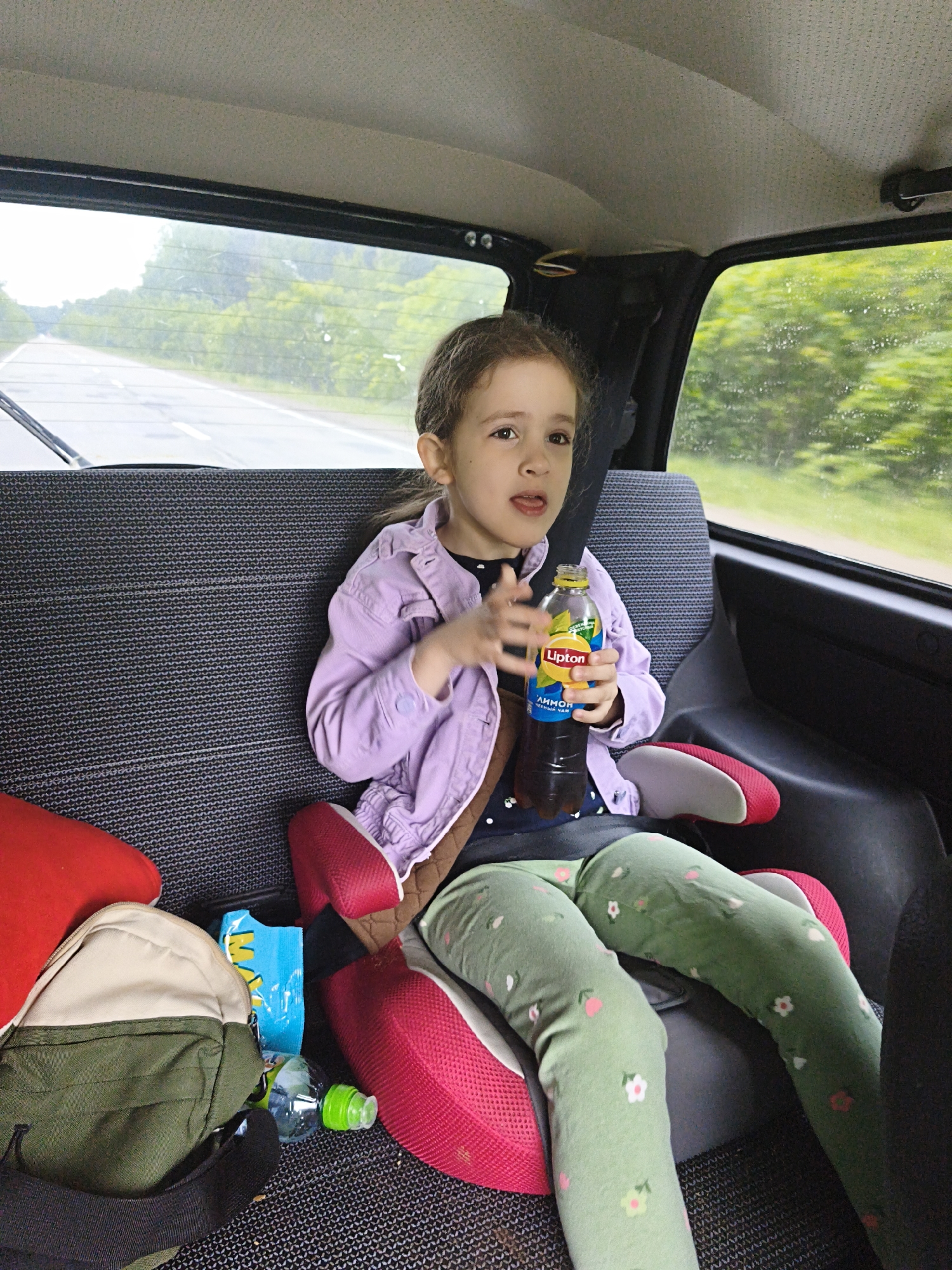A young girl is seated in the back of a car, buckled into a booster seat. She wears a light purple jacket over a dark shirt and green leggings with small floral patterns. In her right hand, she holds a bottle of Lipton iced tea, with the label visible showing "Lipton" and "ЛИПТОН ЧЁРНЫЙ ЧАЙ" (Lipton Black Tea). Her left hand is also near the bottle, fingers slightly blurred as if in motion. Her expression is neutral, with her tongue slightly visible.
The car's interior appears somewhat dated, with a textured grey headliner and fabric car seats. Through the rear window, a road is visible, with green trees lining its sides, suggesting travel outside an urban area. There are visible streaks on the windows, possibly from rain. On the seat next to the girl, a green and beige backpack, a small blue snack bag, and a clear water bottle with a green cap are scattered.
The setting is inside a moving vehicle, likely a Niva in Russia, during the daytime, possibly overcast or after rain. The activity is that of a passenger on a journey.