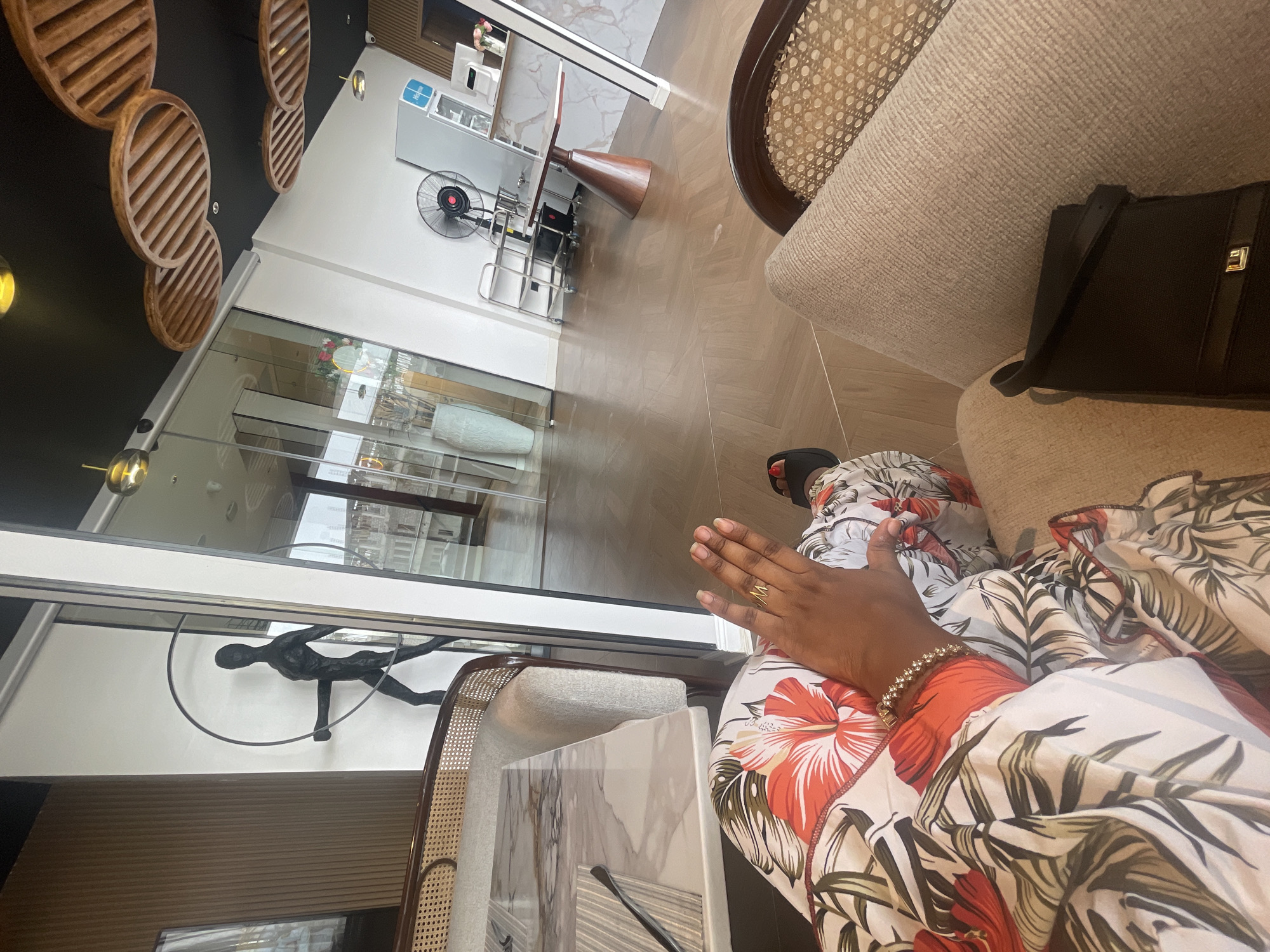 The image shows an interior space, possibly a lounge or waiting area, in what is likely a commercial establishment.
**People:** A person is seated, partially visible from the knees up, wearing a colorful floral-patterned garment. The person has dark skin and is wearing jewelry - a ring and a bracelet.
**Objects:** There is furniture: chairs, possibly sofas. A black purse is visible on the chair beside the person. Art objects and decorations are present: A sculpture of a figure is mounted on the wall, and decorative wooden circles are placed on the ceiling. A large vase is behind a glass partition. A fan and some tables are also visible.
**Environment:** The space appears clean and modern, with large glass windows. The lighting is bright, indicating a well-lit interior. The reflections in the glass hint at an exterior environment, but details are unclear.
**Location:** While the image is said to be from Abuja, Nigeria, it is impossible to confirm the specific location within the city based solely on the visual information provided. The presence of modern decor and commercial furniture suggests a higher-end business or hospitality setting.