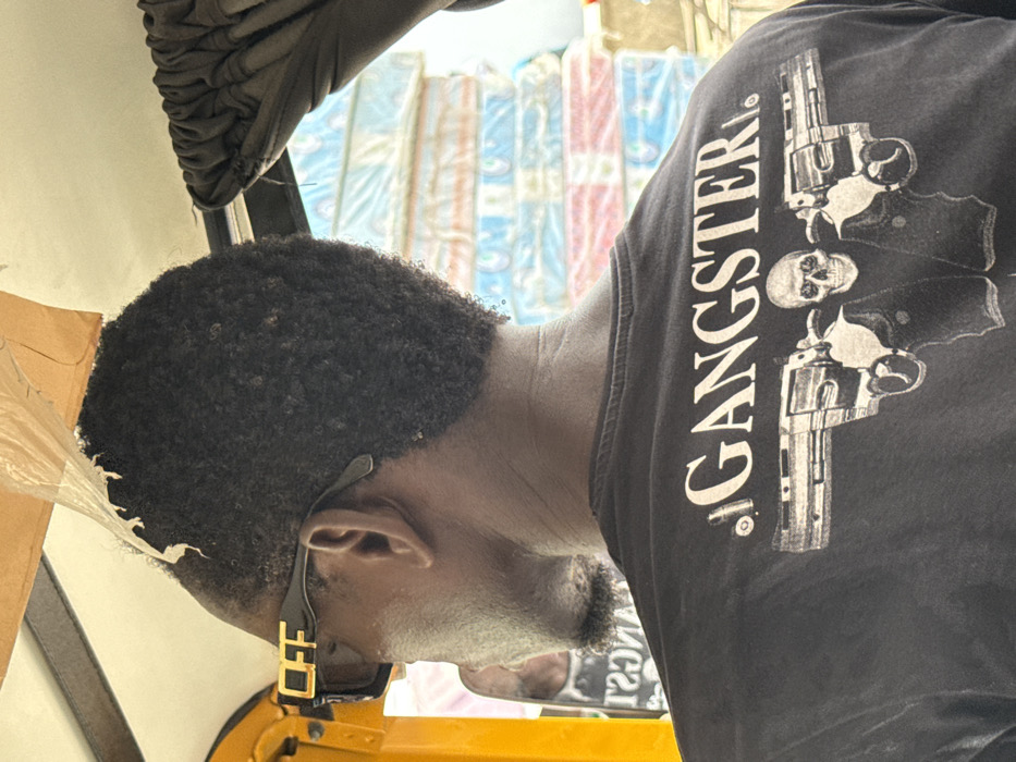 The image presents a close-up, rear-facing perspective of an individual inside a vehicle, likely a keke napep (tricycle taxi), in a bustling urban environment consistent with a market or commercial street in Lagos, Nigeria.
The primary subject is an adult viewed from the back and partial right profile. The individual has dark skin and short, dark, curly hair. They are wearing black sunglasses with a gold "OFF" logo prominently displayed on the right temple. The subject's attire includes a black t-shirt featuring a white graphic design on the back. This design consists of the word "GANGSTER" in capital letters, beneath which two revolvers are depicted, positioned outwards from a central skull wearing a jacket. A spent bullet casing is visible near the letter "G" in "GANGSTER".
Within the vehicle's interior, a yellow structural frame is visible on the left side, and a torn piece of light brown plastic or fabric hangs from the ceiling near the subject's head. A dark, pleated curtain or fabric is present on the right side of the frame.
The background, viewed through the vehicle's opening, is somewhat blurred but reveals key environmental details. A large, tall stack of multiple plastic-wrapped items, identifiable as mattresses or foam cushions, is prominently displayed. These items are horizontally stacked and show various colors, including blue, white, red, and orange, suggesting a retail or wholesale outdoor setup. A blurry reflection of another person wearing a dark shirt with white lettering is faintly visible on the far left. The overall scene depicts typical street activity and commerce found in Lagos.
