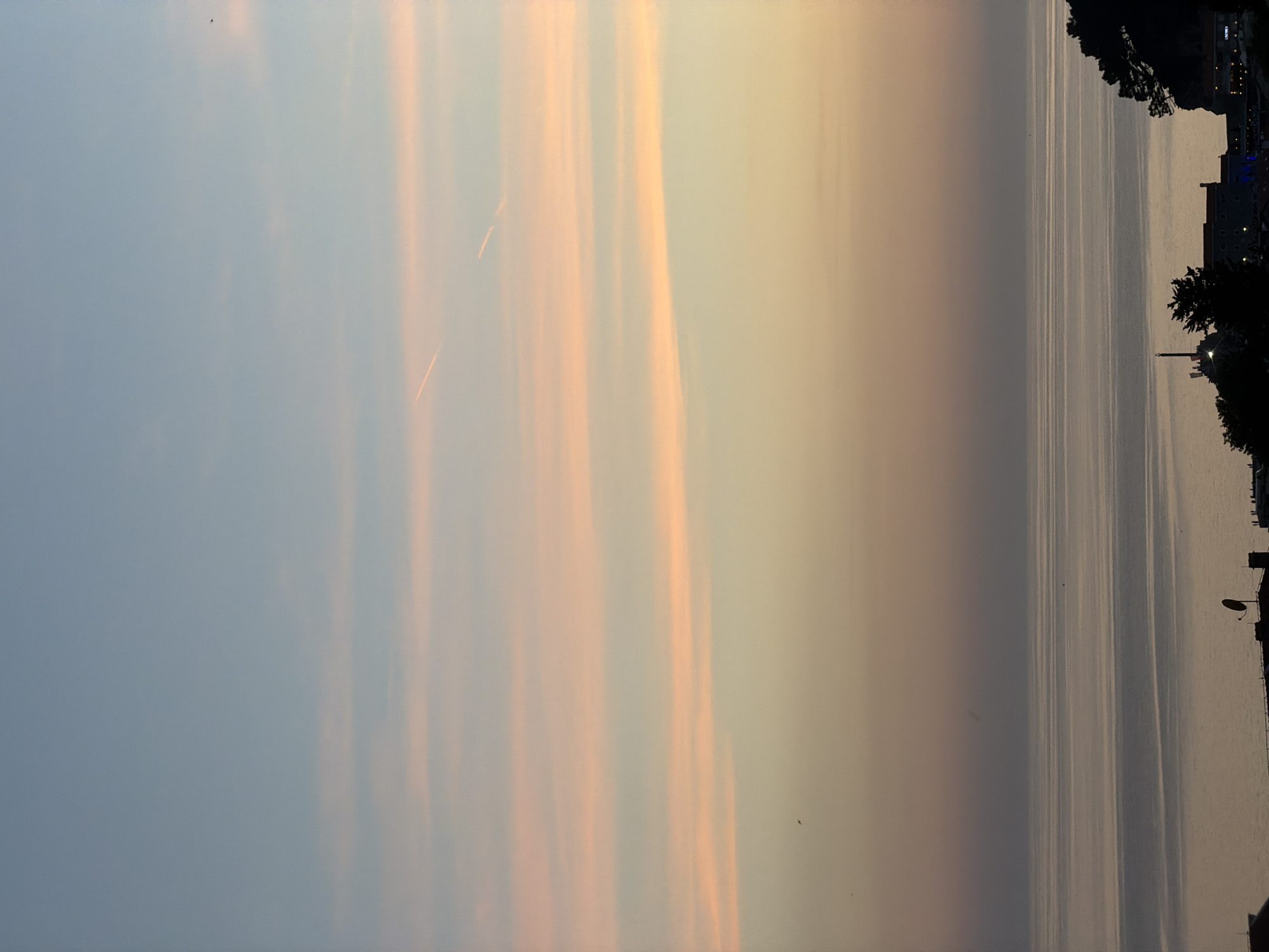 The image captures a sunset over the Adriatic Sea in Petrovac na Moru, Montenegro. The sky transitions from a pale blue at the top to soft hues of pink and orange, indicating the setting sun. Faint horizontal clouds are visible, some tinged with the sunset colors. A contrail from an aircraft streaks across the upper portion of the sky. The calm sea below reflects the muted colors of the sky. In the foreground, silhouetted against the fading light, is a coastal landscape featuring a dark, tree-covered promontory on the right. A cluster of buildings, possibly part of the town, is visible along the shoreline, with structures and antennas silhouetted. The overall scene is serene and atmospheric, characteristic of a coastal sunset.