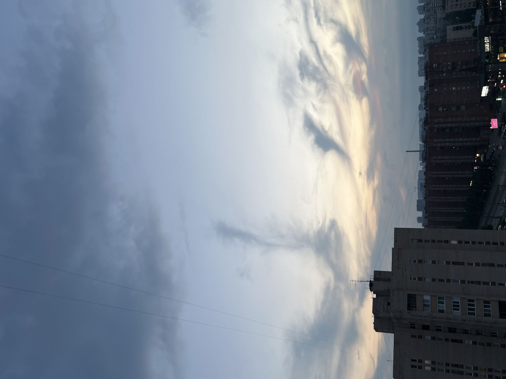 The image captures an urban skyline at dusk in an unspecified location in Russia. The foreground features the upper portion of a light-colored, multi-story brick building with numerous windows. Extending from this building are power lines that traverse the sky. In the midground, rows of darker, brick apartment buildings dominate the scene, with lights visible in some windows. Further into the distance, a dense cityscape of residential buildings stretches across the horizon. The sky above is a mix of dark blue and lighter hues, with wispy clouds tinged with pink and yellow, suggesting the setting sun. The overall impression is one of a vast, densely populated urban area under a dramatic twilight sky.