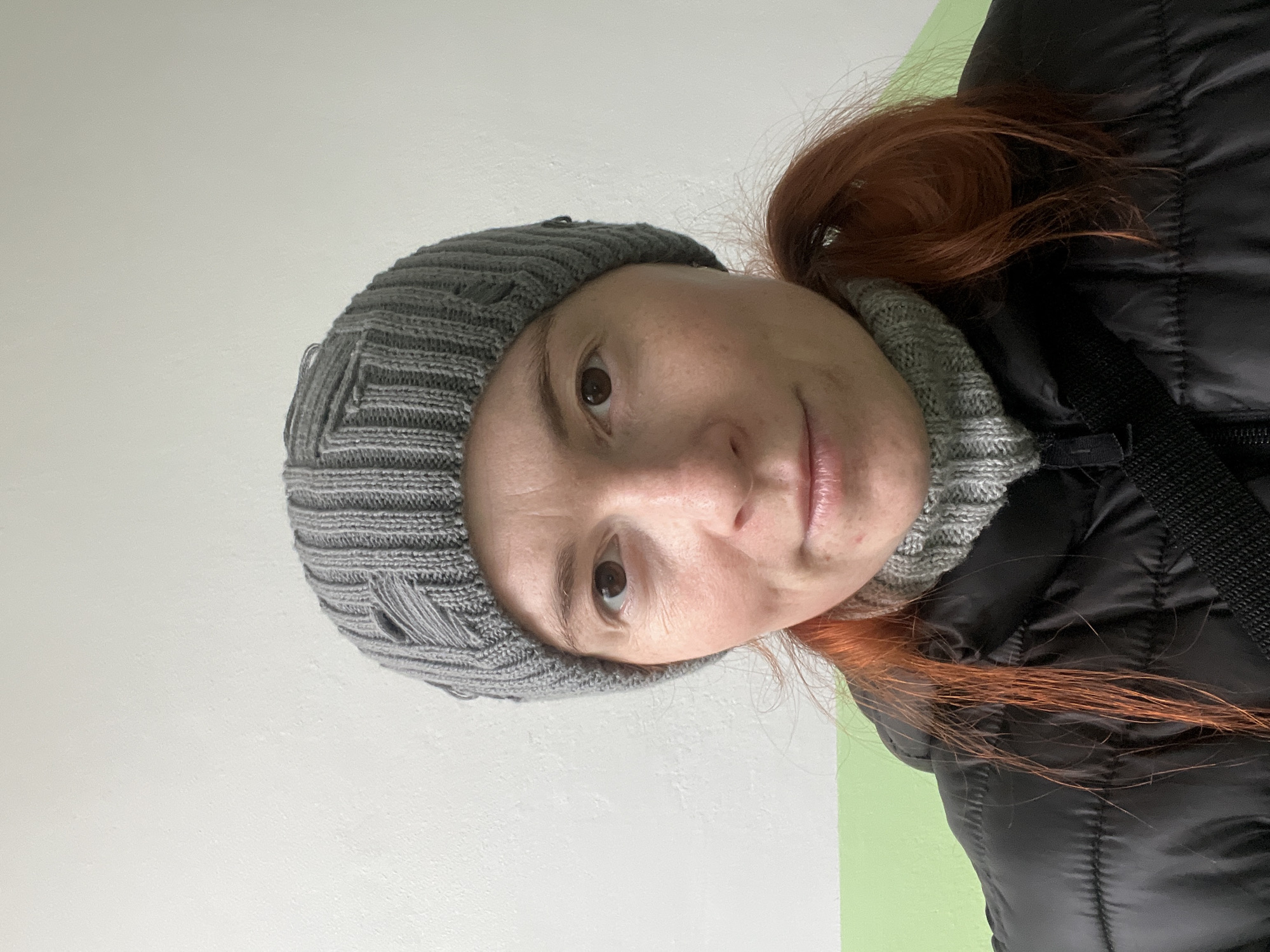 A woman with reddish-brown hair and freckles is wearing a grey knitted beanie and a grey turtleneck sweater under a black puffer jacket. The background appears to be a wall with a split color scheme: a lighter shade on the left and a slightly greener shade on the right. The image is a selfie-style portrait. The location in the city of Chaikovsky, Russia is unknown.