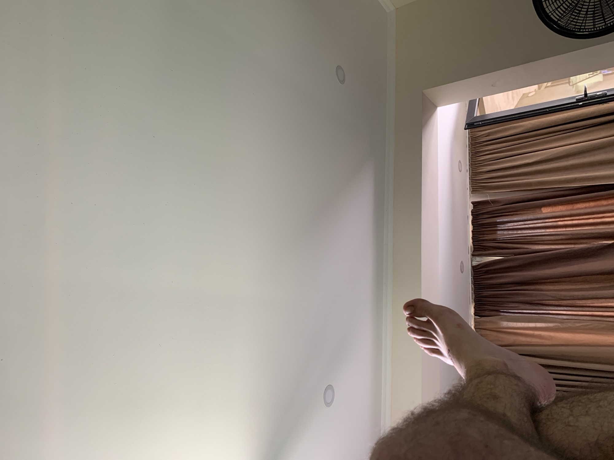 The image depicts a low-angle shot focusing on a person's hairy leg and foot, which is elevated. The foot is in the foreground and positioned on the right side of the frame, with visible toes and toenails. The leg, covered in dark hair, extends from the left side of the image. The background features a white ceiling with two recessed circular lights. Below the ceiling, there is a section of wall painted a light color, topped by white crown molding. Further down, a pair of brown curtains are visible, partially obscuring a doorway or opening. A black fan is partially visible on the far right edge of the frame. The scene appears to be indoors. There is no information present in the image that can be used to determine its location in Kakhati, Georgia.