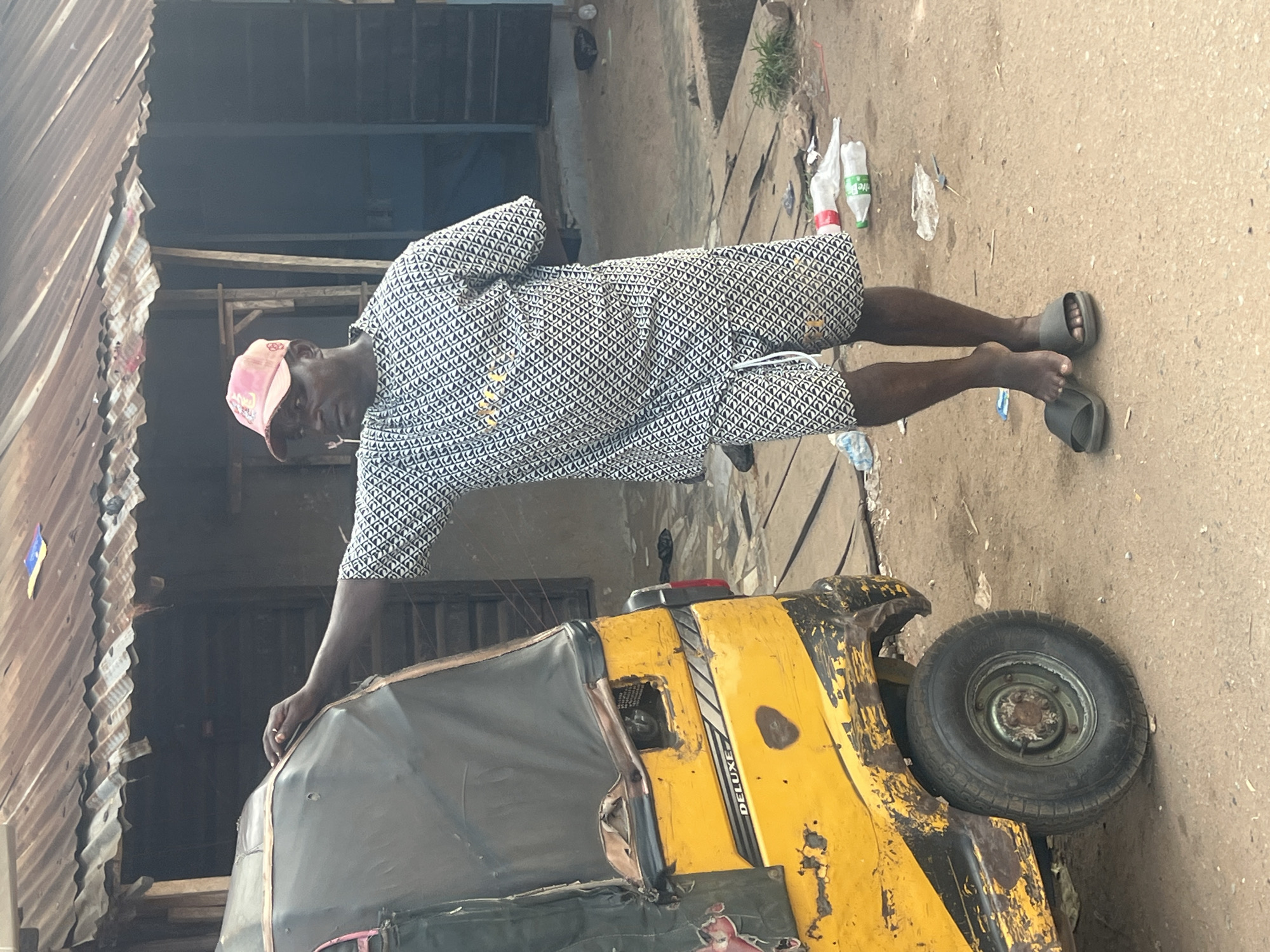 A full-body shot captures a man standing outdoors next to a yellow tricycle in Obosi, Nigeria. The man, with dark skin, wears a short-sleeved, patterned white and black shirt with matching shorts, a pink baseball cap, and dark slides or sandals. He looks directly at the camera with a neutral expression, his right arm resting on the black canopy of the tricycle.
The yellow tricycle, commonly known as a Keke Napep, shows signs of wear and tear, with chipped paint, dark streaks, and some rust. The word "DELUXE" is visible on its side. A black fabric canopy covers part of the passenger area. One tire is clearly visible on the right side of the tricycle.
The scene appears to be an urban or semi-urban setting. The ground beneath the man and the tricycle is a dusty, light brown dirt or pavement. To the left, corrugated metal sheeting forms part of a structure, possibly a building or fence. The background is somewhat dark and indistinct, with hints of other structures and scaffolding. Scattered litter, including empty plastic bottles and wrappers, is visible on the ground near the man's feet and in the background. The lighting suggests it is daytime, possibly under an overcast sky or in a shaded area.