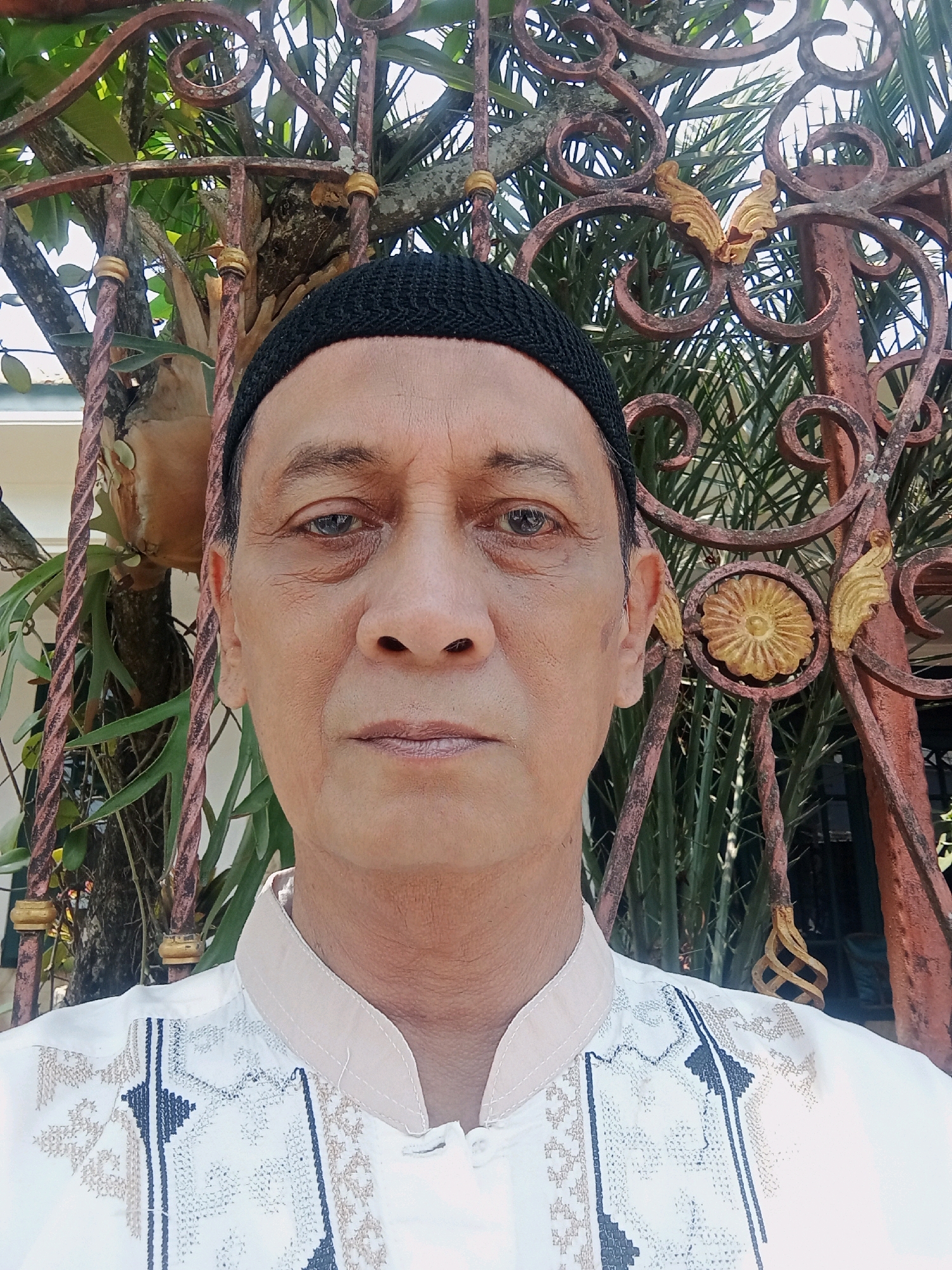 A man wearing a black skullcap and a white, embroidered shirt stands in front of a decorative, reddish-brown metal fence. Behind the fence, lush green foliage is visible. The man's face is turned towards the camera, and his expression is neutral. The image appears to be a selfie, taken outdoors during the day. The presence of ornate metalwork and tropical vegetation suggests a location in a warm climate, possibly in the city of Malang, Indonesia, known for its pleasant climate and cultural heritage. The details of the man's attire, particularly the intricate embroidery on his shirt, could indicate traditional Indonesian clothing. The overall scene is one of a man posing for a photograph in a garden or courtyard setting.