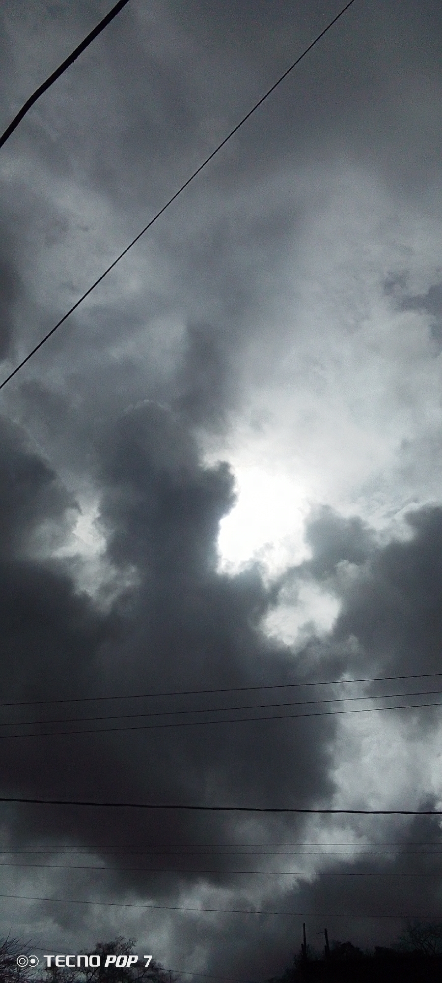 The image displays an overcast sky over Altagracia, Venezuela, characterized by dense, dark grey cumulonimbus-like cloud formations. A single, intense white light source, presumed to be the sun, is visible through a substantial opening in the central cloud mass, creating a dramatic contrast. A prominent, thick black cable or power line traverses diagonally from the upper-left corner towards the mid-right. Another thinner, dark cable runs from the upper-left towards the upper-right. Below these, a cluster of approximately six horizontal utility lines spans across the lower half of the frame. The bottom edge of the image shows a very dark, indistinct area, suggestive of silhouetted treetops or distant structures, with two vertical utility poles faintly visible on the lower-right. No human figures, animals, or explicit actions are present. A digital watermark "© TECNO POP 7" is inscribed in the lower-left corner.
