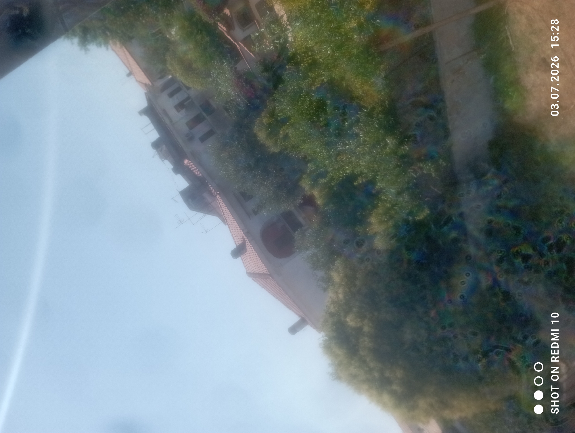 The image, taken from a slightly elevated angle, shows a view of a building with a red-tiled roof and multiple windows, partially obscured by lush green trees and foliage. The sky is bright blue and appears slightly hazy. The overall scene suggests an outdoor setting in a residential area. The bottom left corner of the image displays the text "SHOT ON REDMI 10", and the bottom right corner shows the date "03.07.2026" and time "15:28". The image has some visual artifacts, including circular distortions and color aberrations, particularly in the lower half. The scene does not provide specific details that can definitively place it in Ardea, Italy, beyond what might be inferred from general architectural styles or vegetation, which are not unique to that location.
