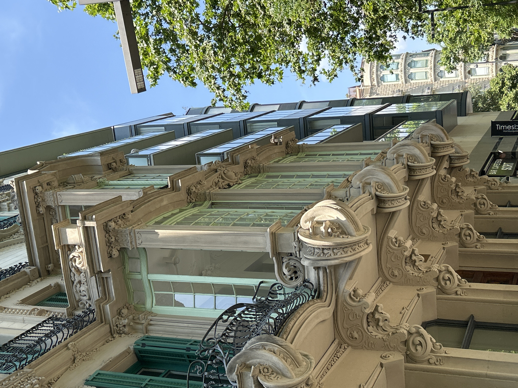 The image shows a low-angle view of a building in Barcelona, Spain. The building has a combination of architectural styles, with an older, ornate facade at the lower levels, featuring detailed carvings, decorative balconies with wrought-iron railings, and window shutters. Above this, there is a modern addition with a series of angular glass windows. The sky is visible in the background, along with the branches of trees that overhang the building. A street light is also visible at the top of the frame. The lighting suggests it is daytime, with the sun illuminating parts of the building. A sign is partially visible on the right side of the building.