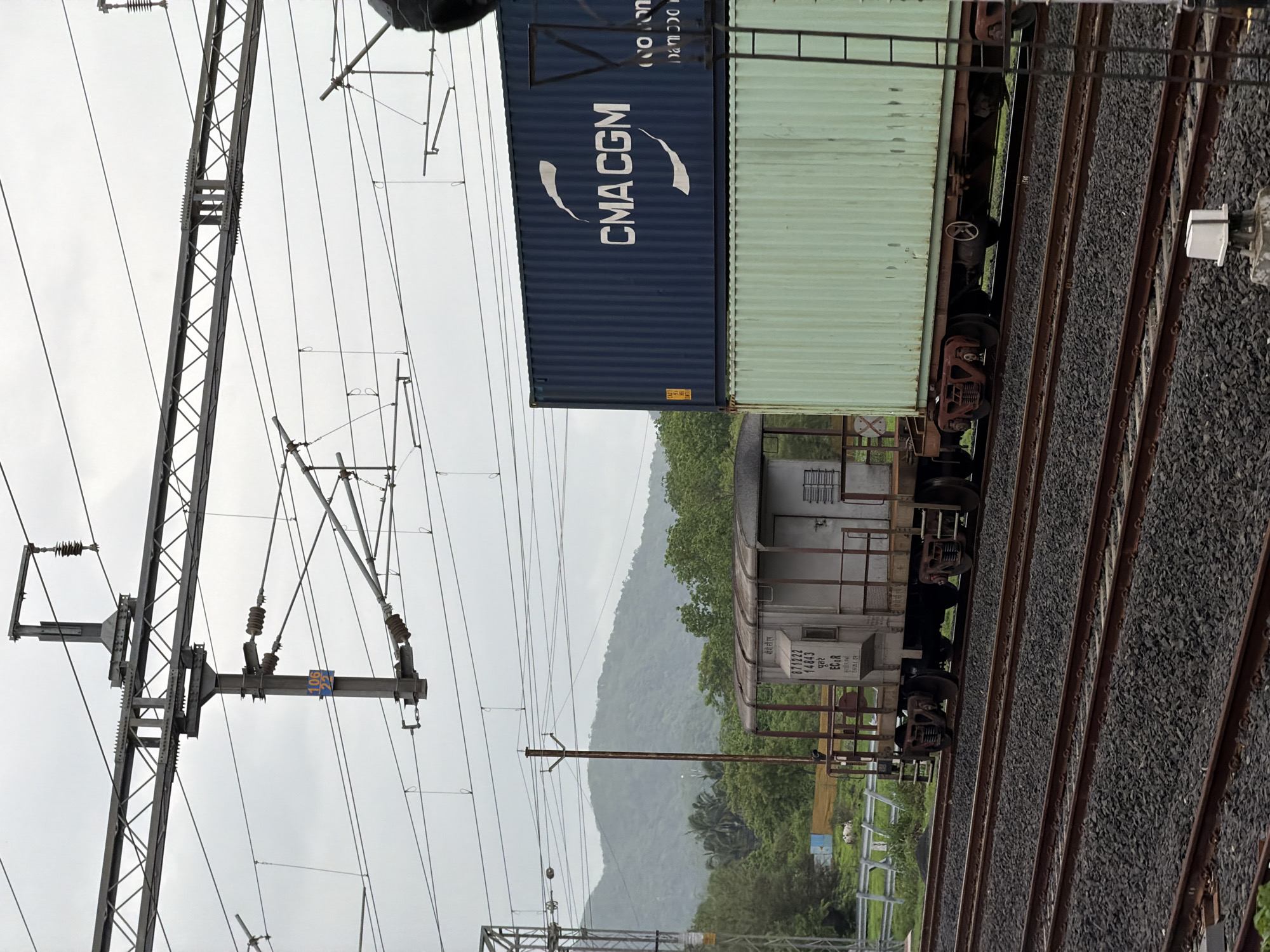 This image depicts a freight train in Saphale, India, moving along a railway track beneath an overhead electrical catenary system. The train consists of multiple wagons. The foremost wagon visible is a grey, open-topped carriage with a visible serial number "871222 14843" and the designation "ECoR". It appears to be carrying a stacked load. On top of this wagon, and extending further back, are two large shipping containers. The upper container is dark blue and prominently displays the logo and text "CMA CGM". The container below it is a pale green color. The railway tracks are laid on a bed of dark ballast. In the background, a lush green, mountainous landscape with trees and foliage is visible under an overcast sky. A metal pole with electrical insulators and wires is prominent in the middle ground, supporting the catenary system. Another metal pole, part of a signal or utility structure, is visible on the right side of the frame.