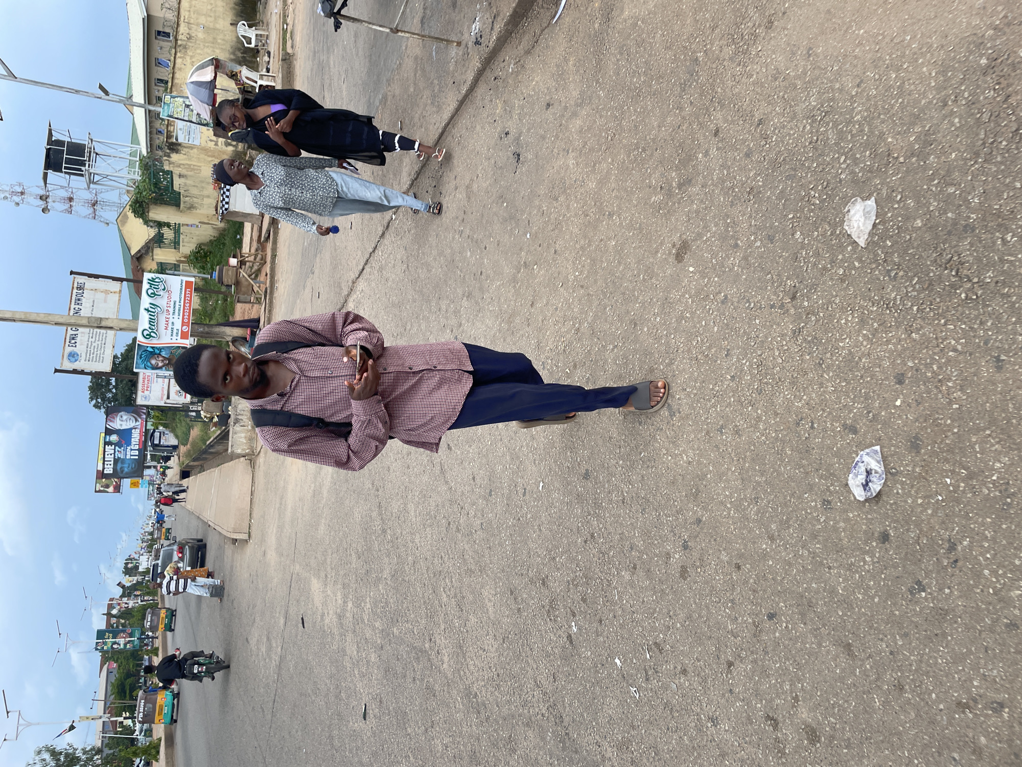 In the city of Jos, Nigeria, on a paved street with sparse vegetation and overhead utility poles, a young man wearing a red and white checkered shirt, dark blue pants, and dark sandals walks towards the camera while looking down at his phone. He carries a black backpack. Behind him, two women walk away from the camera; one in a white floral-patterned shirt and light blue pants, the other in a black outfit. A motorcycle with a rider is visible further down the street, along with several other vehicles and pedestrians. Signs with text and images are posted on buildings along the sidewalk. The ground is littered with pieces of trash. The sky is cloudy.