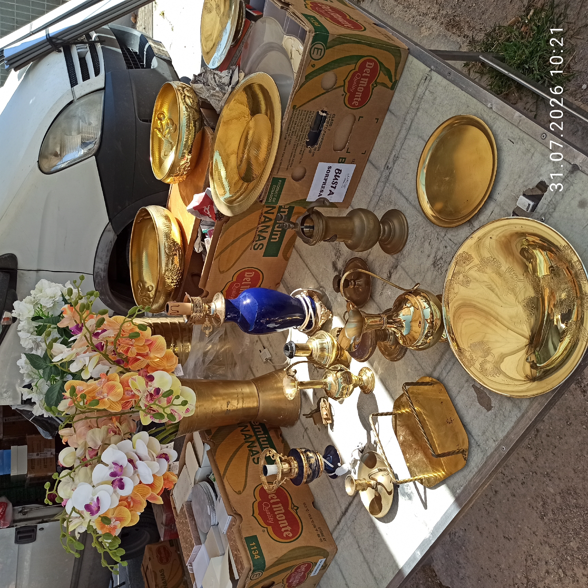This is a street-level photograph taken at an outdoor market in Modugno, Italy, on July 31, 2026, at 10:21 AM. The image features a table laden with various items, predominantly gold-colored decorative and functional objects, set up in front of a white van. The van is partially visible, with its front bumper, headlight, and grille in the upper right and left portions of the image. In the background, stacked boxes, including a "Del Monte" branded box of bananas, are visible.
The foreground is dominated by the display table. On the table, there is a large arrangement of artificial orchids in shades of white, peach, and orange, displayed in a tall gold vase. Scattered among the orchids and other items are several brass or gold-plated decorative pieces. These include a hammered gold bowl, an oval gold dish with an embossed floral pattern, and a round gold plate. There are also several smaller brass items such as a candlestick, a small pitcher or jug with a handle, and an ornate blue and gold table lamp. A gold metal napkin holder with twisted wire handles is positioned on the table, along with a small gold tray. Some of the items are casting distinct shadows, indicating bright sunlight. The table surface appears to be made of a marble-like material. The ground around the table is a mix of concrete and soil with patches of grass. No people are visible in the photograph.