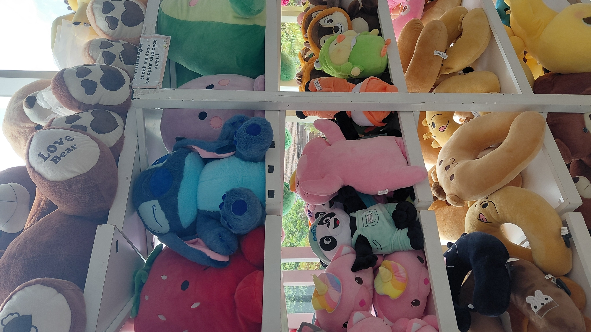 The image shows a retail display of stuffed animals in Purwokerto, Indonesia. The stuffed animals are arranged on white shelves. In the top shelf, there are large teddy bears. The middle shelf features a variety of plush toys including Stitch, a strawberry, a pink elephant, a panda in overalls, a pink pig, and some green and orange plush toys. The bottom shelf displays several U-shaped neck pillows, some resembling animals, and other assorted plush items. A small sign with Indonesian text, "Sudah menjaga kerapian display kami :)" which translates to "Already keeping our display tidy :)" is visible on the middle shelf. The background suggests an indoor setting with natural light coming from windows, revealing some greenery outside, possibly indicating a shop or a store.
