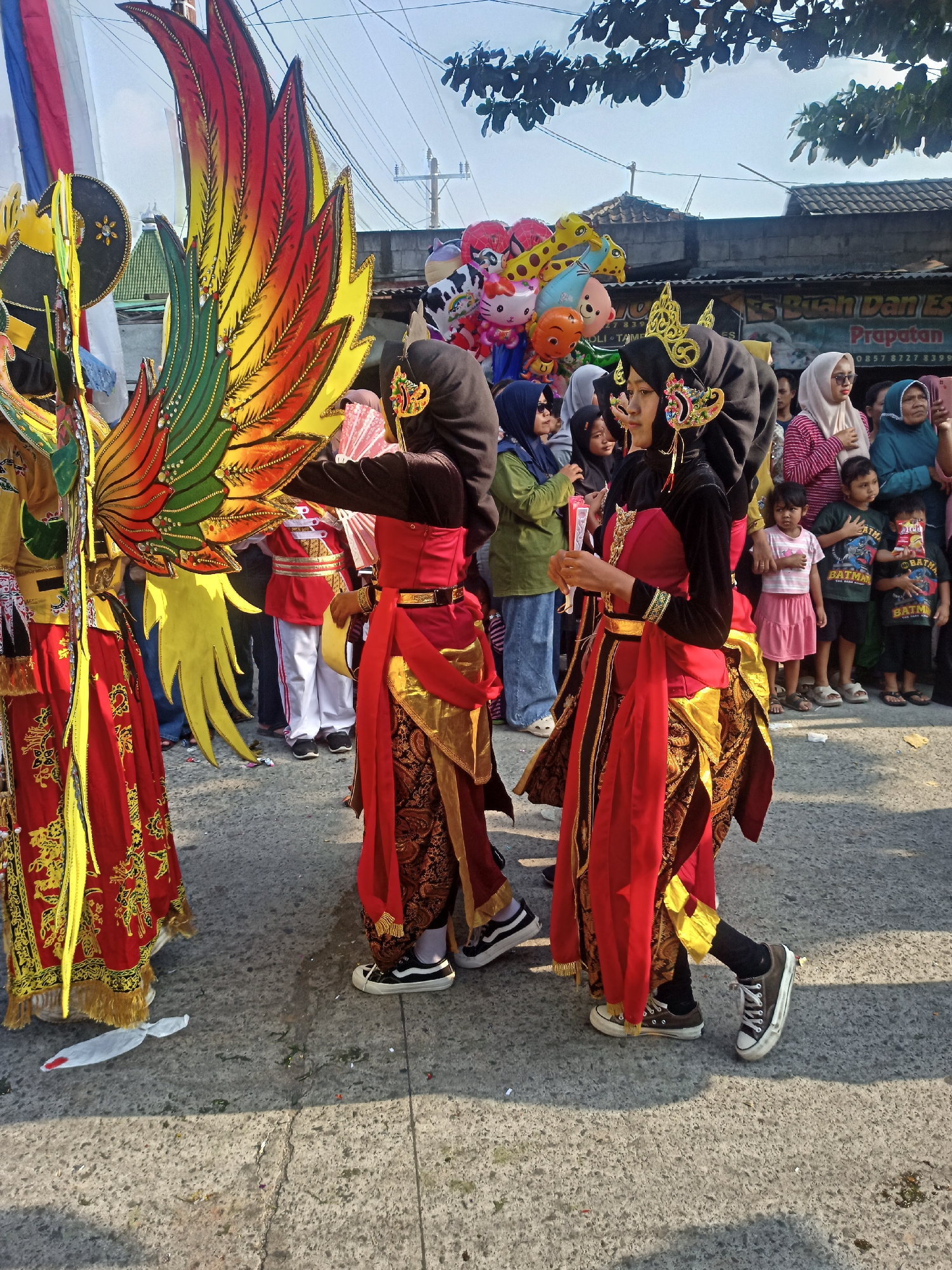 A group of young women in traditional Indonesian costumes are participating in an outdoor parade in Saren, Indonesia. The women are adorned in vibrant red and gold garments, featuring intricate patterns and gold headpieces. Some of them are wearing black hijabs. One individual is holding a fan, while another is carrying a large, colorful wing-like costume, likely part of a parade float or performance. In the background, a crowd of spectators, including children and adults, are gathered to watch the event. Balloons, including animal-shaped ones like a giraffe and a pig, are visible in the background, adding to the festive atmosphere. The scene takes place on a paved street under a bright, sunny sky.