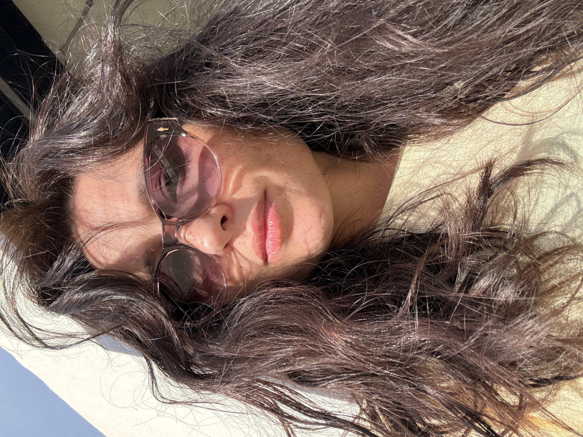 A close-up, tilted portrait features a person, likely female, lying down outdoors under strong, direct sunlight. The person's long, dark, wavy hair is spread out around their head, catching the light with numerous individual strands and highlights visible.
The subject wears fashionable sunglasses with pink-purple tinted lenses, which obscure most of their eyes and cast shadows across the upper part of their face. A subtle smile is visible on their lips, and their skin is illuminated by the bright sun, showing texture and some small moles or freckles.
The strong overhead or angled sunlight creates a high-contrast scene, with bright areas on the hair and skin, and distinct shadows cast by the sunglasses and hair onto the face and the surface below. The person appears to be resting on a light-colored, possibly cream or white, textured surface, which could be fabric or a lounge cushion.
The bright lighting and relaxed pose suggest a sunny daytime setting, consistent with the location context of Safaga, Egypt, implying good weather and an outdoor leisure activity like sunbathing or resting. There is no visible text in the image.