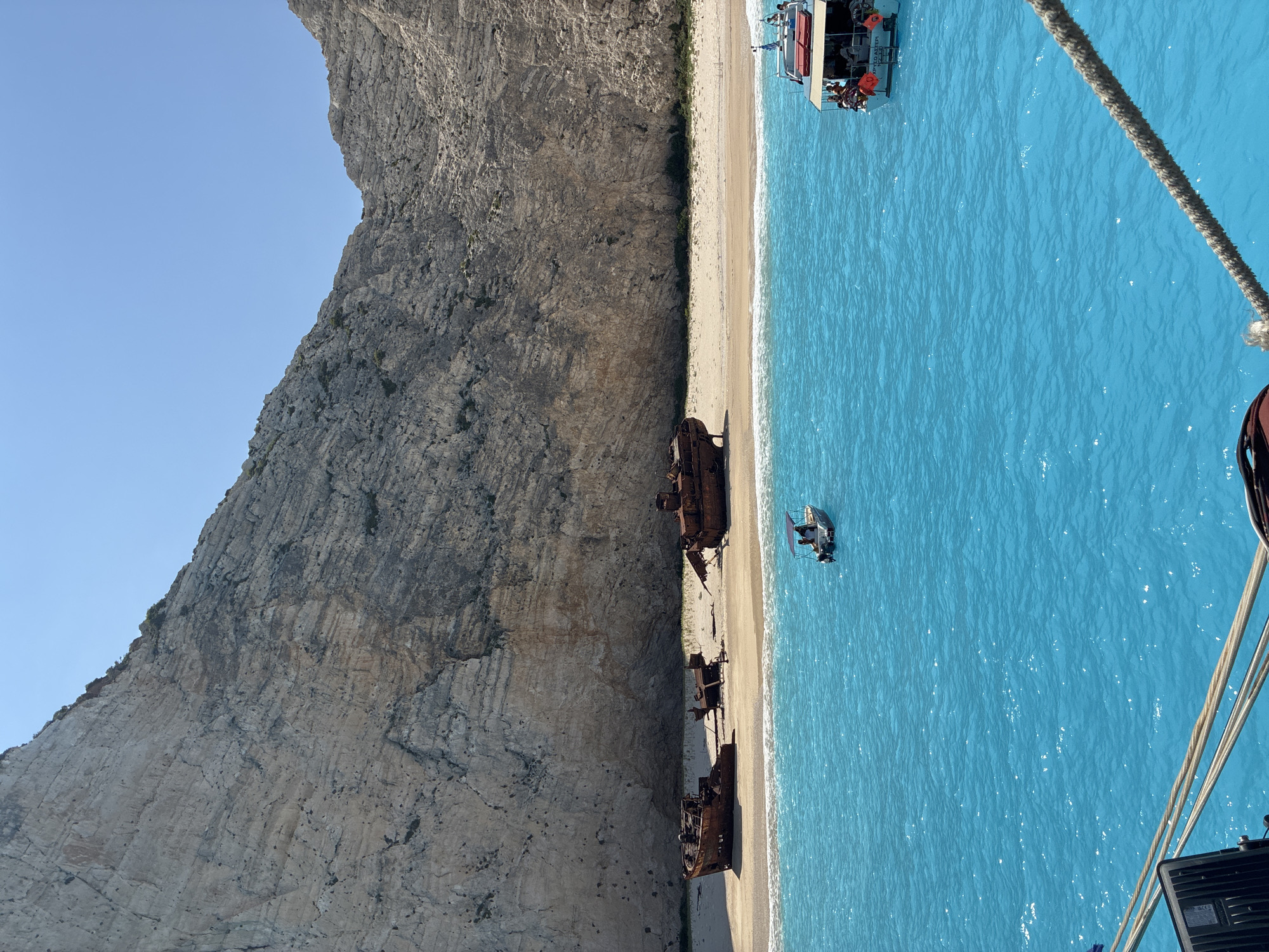 The image depicts a scenic view of the Shipwreck Beach (Navagio) in Zakynthos, Greece, with the iconic rusty shipwreck visible on the white sandy shore. The turquoise waters of the Ionian Sea are calm and inviting, reflecting the clear blue sky above. The backdrop is dominated by towering limestone cliffs, characteristic of the Navagio landscape. Two boats are present in the water: a small dinghy with two people onboard in the middle ground, and a larger tour boat with several passengers in the foreground, to the right. The tour boat has a Greek flag and the name "XPYSO ALTEPI" written on its stern. The foreground also shows a rope and part of another vessel, suggesting the viewpoint is from a boat. The scene is bathed in bright sunlight, creating a warm and inviting atmosphere.