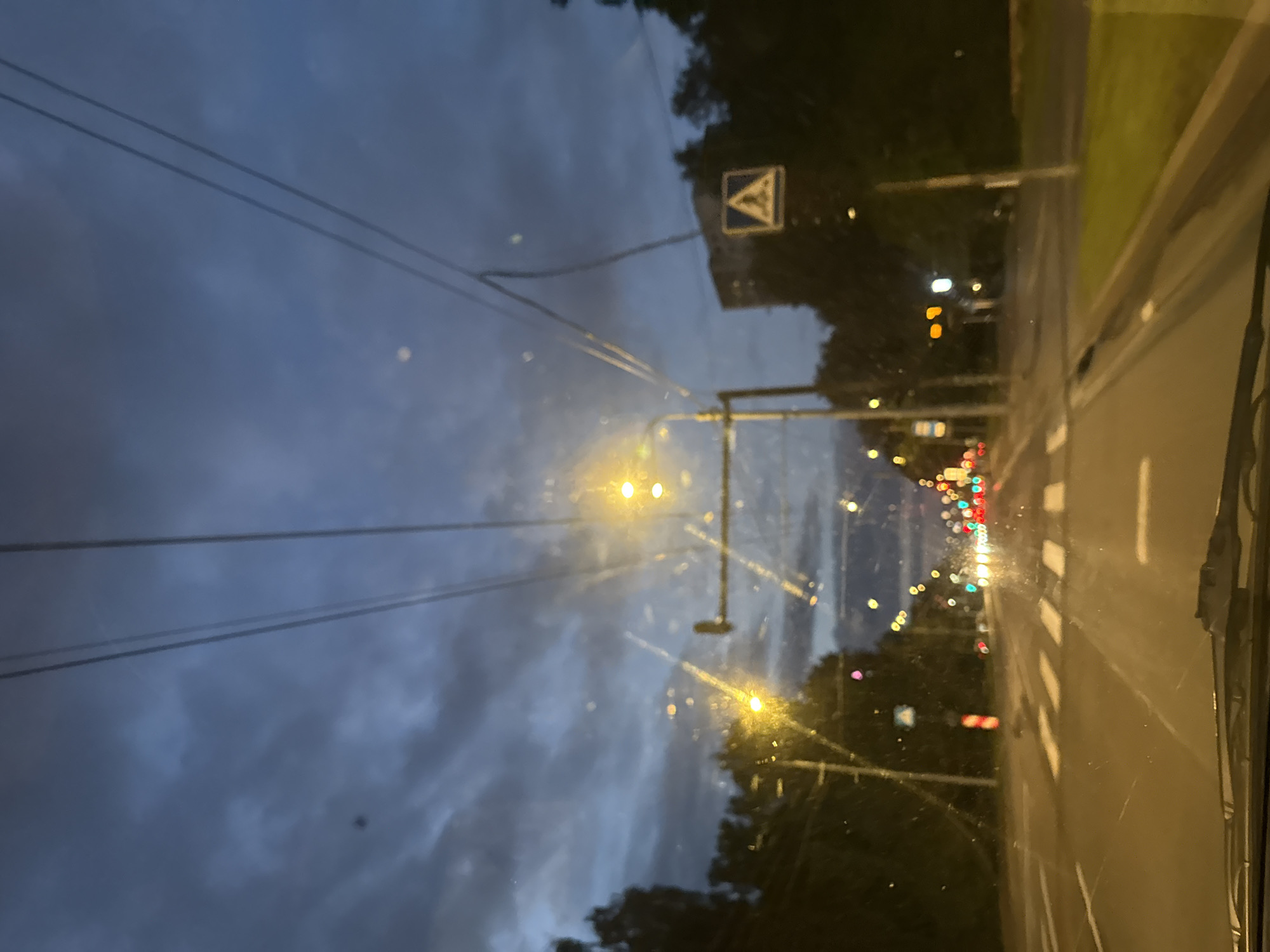 The image shows a view from a vehicle traveling down a street in Tallinn, Estonia, at dusk. The sky is dark blue with heavy clouds, and streetlights are illuminated, casting a warm glow on the wet road and a crosswalk. Trees line the sides of the road, and a few buildings are visible in the distance. A pedestrian crossing sign is also present. The windshield of the vehicle is visible in the foreground, showing some streaks and smudges.