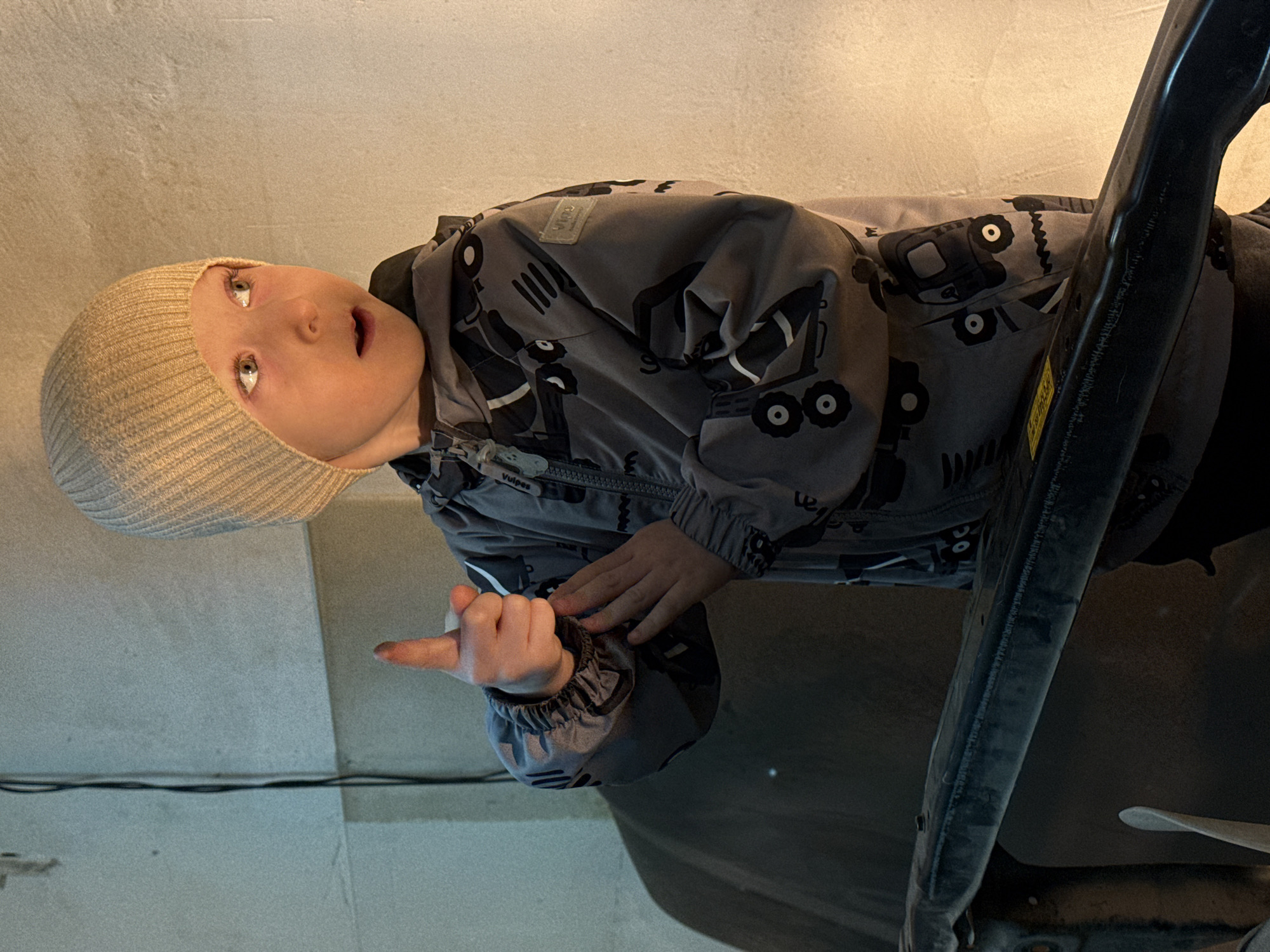A child wearing a light tan beanie and a gray jacket with black tractor designs is pointing with their right hand. The child appears to be indoors, with a light-colored wall visible in the background. A black metal object is partially visible on the right side of the image. The location is not identifiable as Perm, Russia based on visual information.