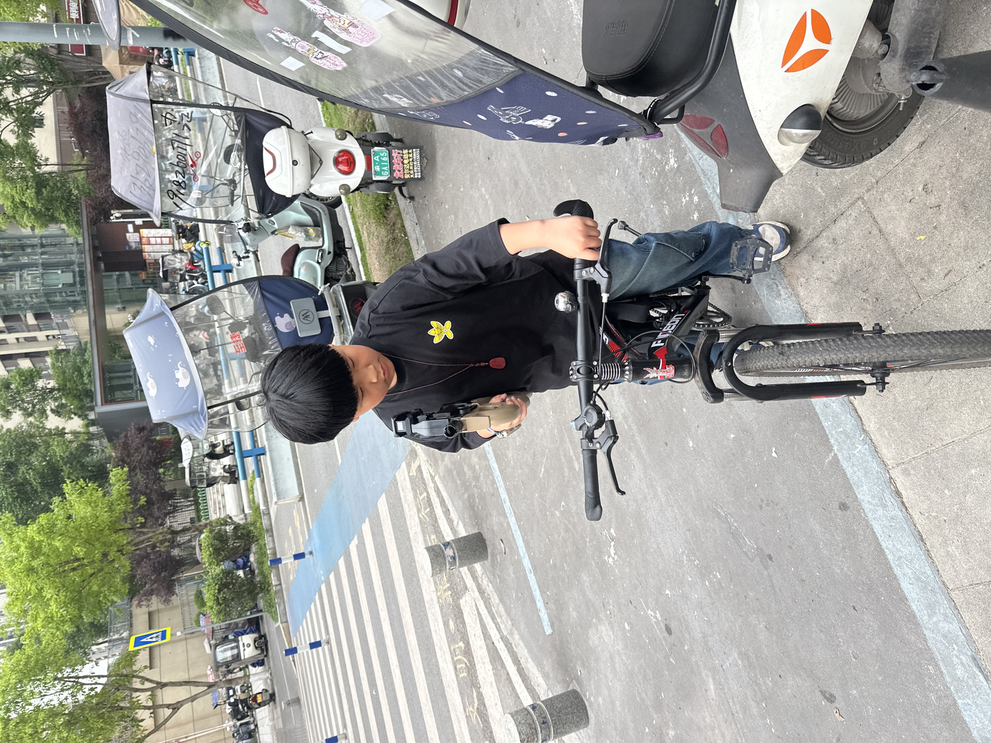 A medium shot depicts a person, presumably a young boy, sitting on a bicycle in an urban environment. He is positioned on a crosswalk marked with white and blue lines, his hands gripping the handlebars. The boy is wearing a black long-sleeved shirt with a yellow floral emblem and blue jeans. He is holding a beige-colored object. The bicycle has black handlebars and a red and black frame, with the word "FIVE" visible on the frame.
The setting is a street in Chongqing High-tech Zone, China. Several electric scooters are parked along the sidewalk, some covered with sunshades or protective canopies. A white scooter with an orange logo and a black seat is parked near the bicycle. The license plate of a white scooter nearby is visible, reading "渝・A GA165." Street signs and trees line the background, adding to the urban context. Buildings are also visible in the distance.