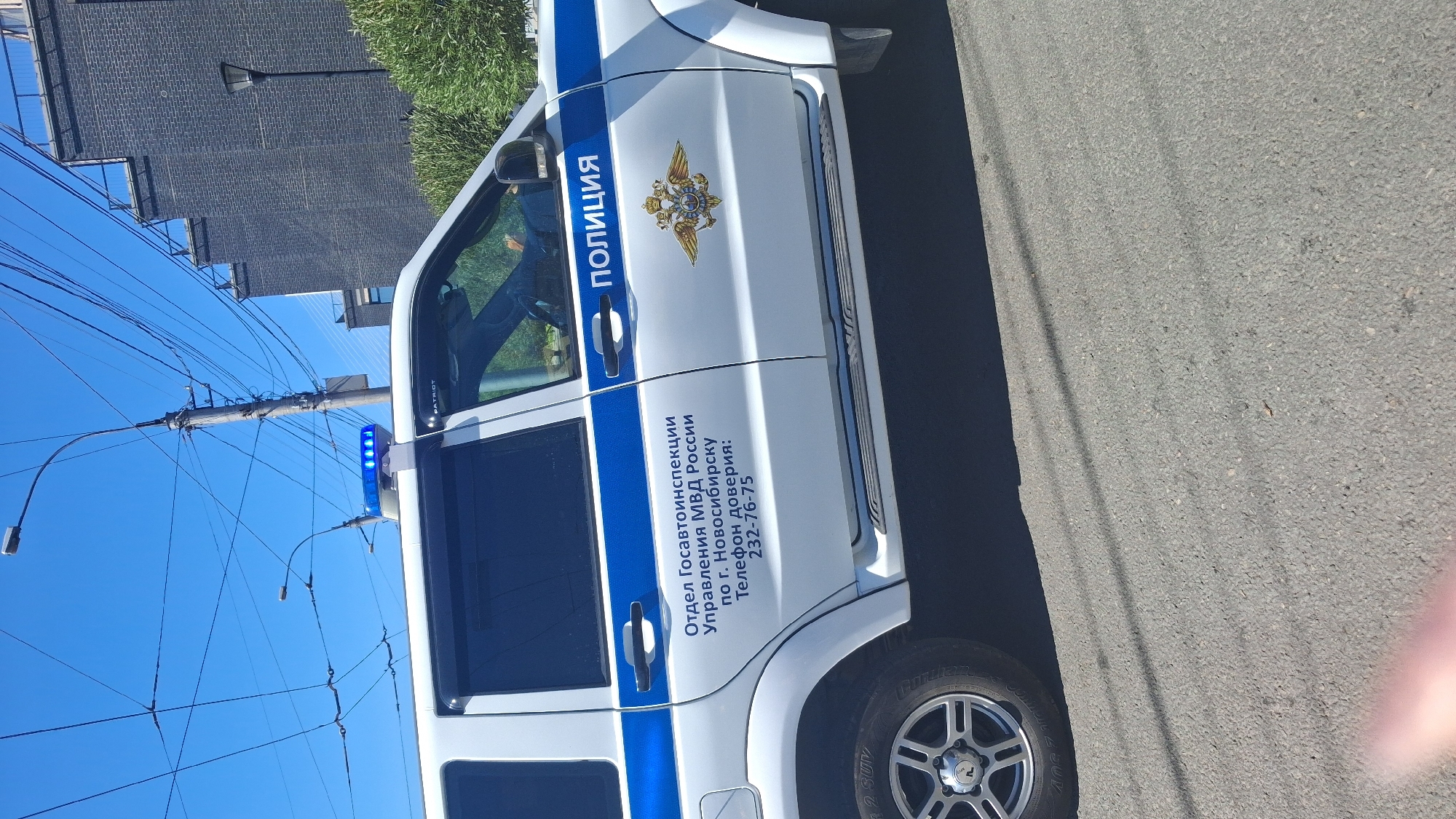 A white SUV with blue stripes and "ПОЛИЦИЯ" (Police) written on its side is parked on an asphalt road under a clear blue sky. The vehicle has a blue flashing light bar on its roof and an emblem on its side. Text on the vehicle reads "Отдел Госавтоинспекции Управления МВД России по г. Новосибирску" (State Road Inspection Department of the Ministry of Internal Affairs of Russia for the city of Novosibirsk) and "Телефон доверия: 232-76-75" (Trust phone: 232-76-75). Overhead wires and a utility pole are visible in the background, along with a dark brick building and green foliage.