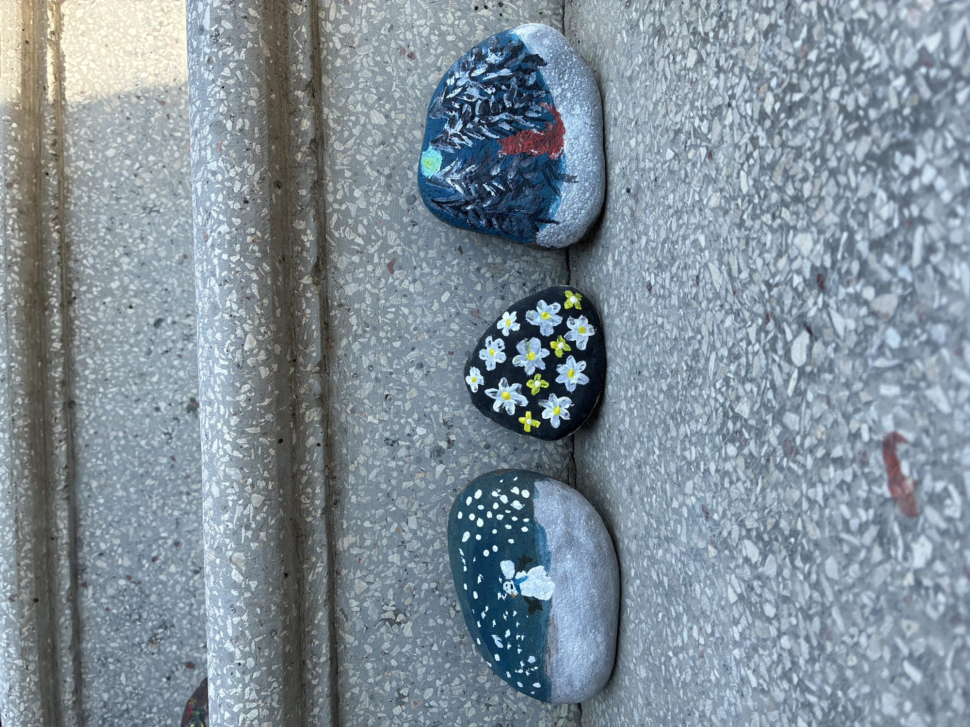 Three painted rocks are arranged on a granite surface, possibly a step or pathway, in Surami, Georgia. The rock on the left depicts a winter scene with a snowman under a starry, snowy sky. The middle rock is painted black and adorned with white and yellow flowers. The rock on the right features a nighttime forest scene with tall trees, a moonlit sky, and a red figure, possibly an animal, in the snow. The granite surface has a rough texture with visible aggregate.