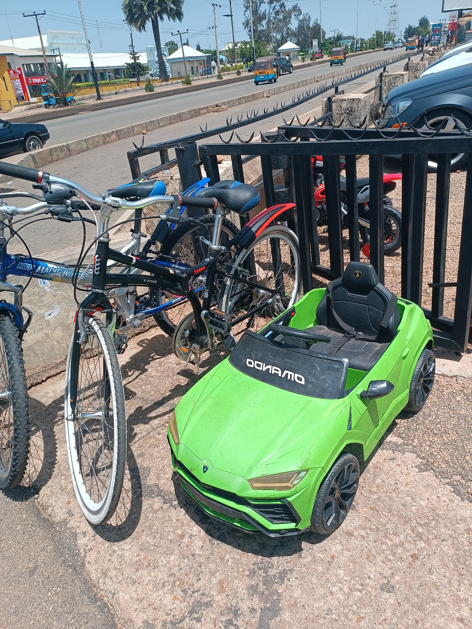 This image depicts a street scene in Jos, Nigeria. In the foreground, a bright green toy car, resembling a Lamborghini, sits on a concrete surface. Beside it, two bicycles are parked. The bicycle on the left has a blue frame, while the one on the right has a black frame. Behind the toy car and bicycles, a black metal fence runs horizontally across the image. Beyond the fence, a busy road is visible with several vehicles, including cars and auto-rickshaws, moving in both directions. The background shows buildings and palm trees, suggesting an urban environment. The sky is clear and blue.