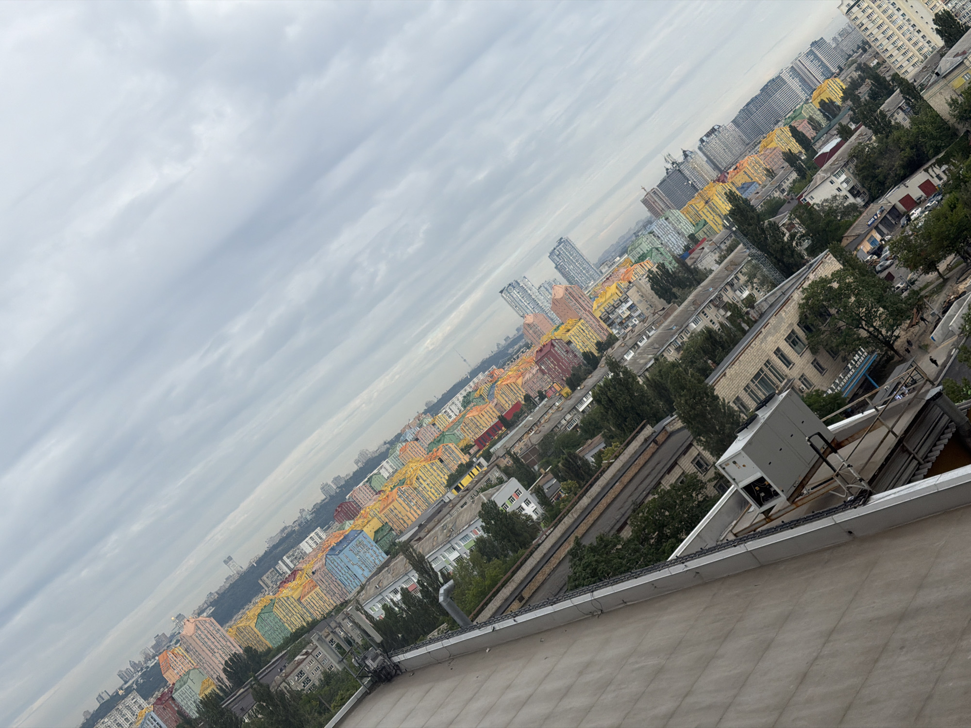 The image shows a high-angle view of a cityscape in Kyiv, Ukraine, with a distinctive row of brightly colored apartment buildings. The buildings are painted in a variety of vibrant colors, including yellow, orange, pink, green, and blue, creating a striking visual effect against the overcast sky. The perspective is tilted, giving a sense of depth and scale to the urban environment. In the foreground, a portion of a building's rooftop is visible, with an air conditioning unit mounted on a metal frame. Beyond this, the colorful buildings form a dense residential area interspersed with trees and other urban structures. Further in the distance, taller, more modern skyscrapers can be seen, along with a hazy horizon characteristic of a large city. The overall scene depicts a residential district within Kyiv, notable for its unique architectural use of color.