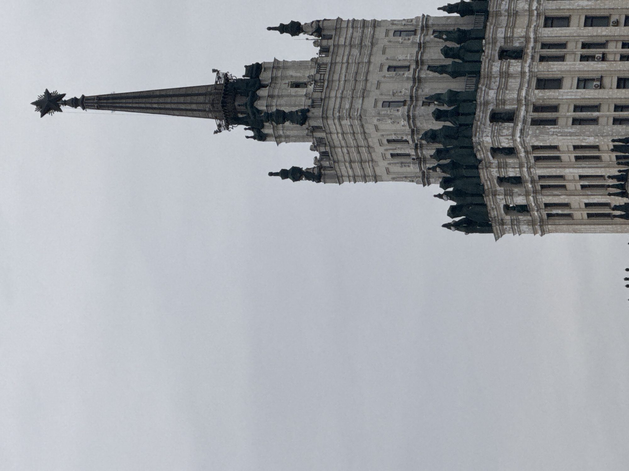 The image shows a building with a prominent spire, crowned with a star. The architectural style appears to be Stalinist, commonly found in Moscow, Russia. The facade of the building is light in color, with multiple floors and a series of windows. The sky is overcast, creating a neutral backdrop. The building is oriented vertically in the image, starting from the top and moving toward the bottom.