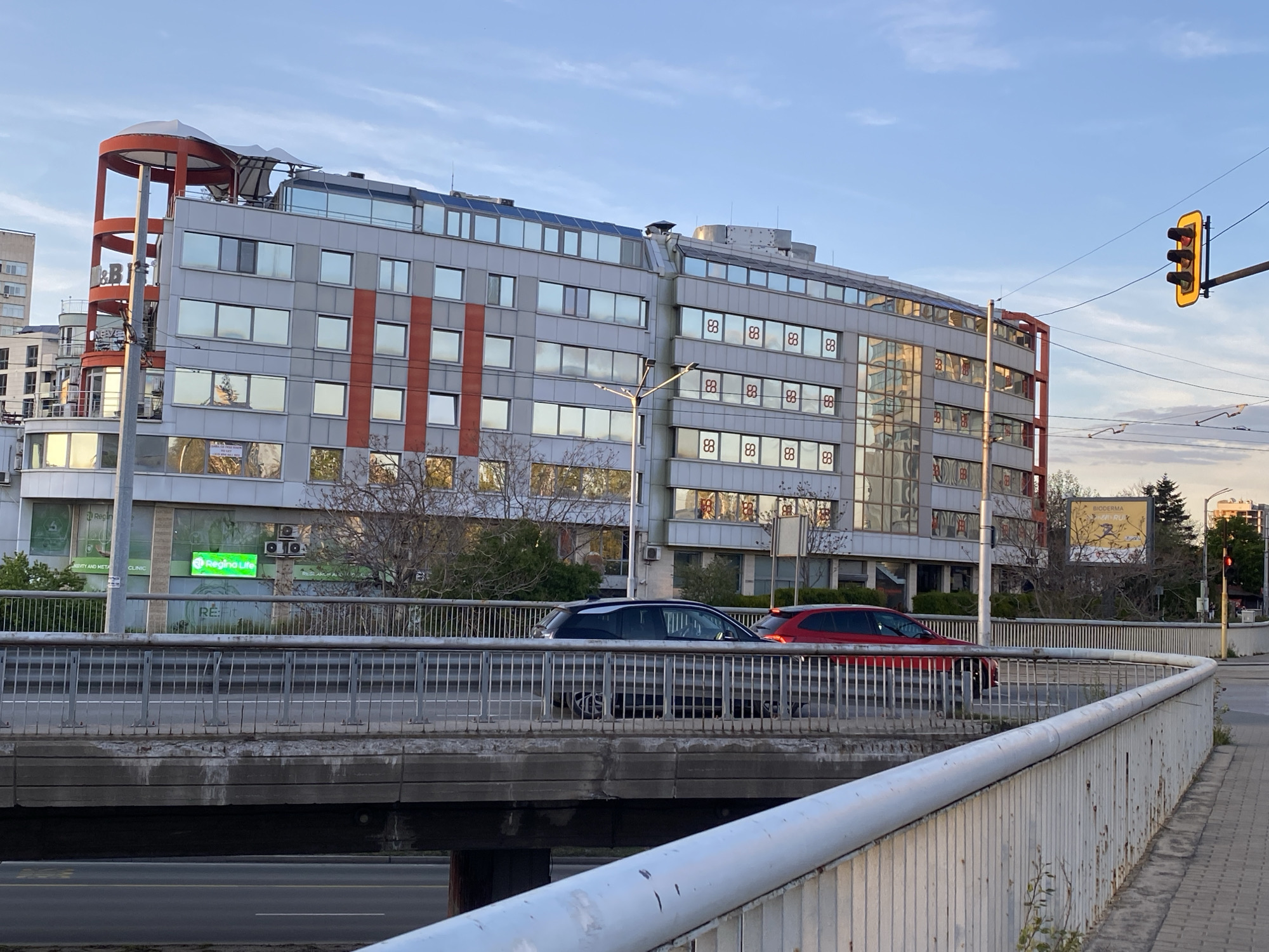 This image captures a late afternoon urban scene in Sofia, Bulgaria, featuring a large, modern building, an overpass, and a street. The time of day is evident from the warm, golden light illuminating the scene and casting long shadows, suggesting either sunrise or sunset, with clear skies and scattered clouds.
The central subject is a multi-story building with a contemporary design, characterized by a grey facade, numerous reflective windows, and distinctive orange-red structural elements. On the left side, a large red, curved structure extends upwards, topped with a white canopy. The building features several visible signs: a prominent green illuminated sign displays "Regina Life" with "CLINIC" below it, and "NEVITY AND META" to its left. Another small green sign reads "RE:fit". Many windows across the building display a repetitive logo resembling two intertwined 'S' shapes or a stylized 'infinity' symbol.
In the midground, an elevated concrete overpass or bridge spans across the view, with a metal railing. Two cars are visible on the bridge: a dark grey/black hatchback (possibly a BMW i3) and a red hatchback. A white metal railing runs along a pedestrian walkway in the foreground on the right.
Further urban elements include a yellow traffic light hanging over the road on the right, and utility poles and wires crisscrossing the sky. In the background, behind the bridge, a large billboard is partially visible with the text "BIODERMA" and "RUR" below it. Trees and shrubs, mostly bare, are scattered around the base of the building and in the background, suggesting late autumn or early spring. The overall atmosphere is that of a typical, busy city environment.