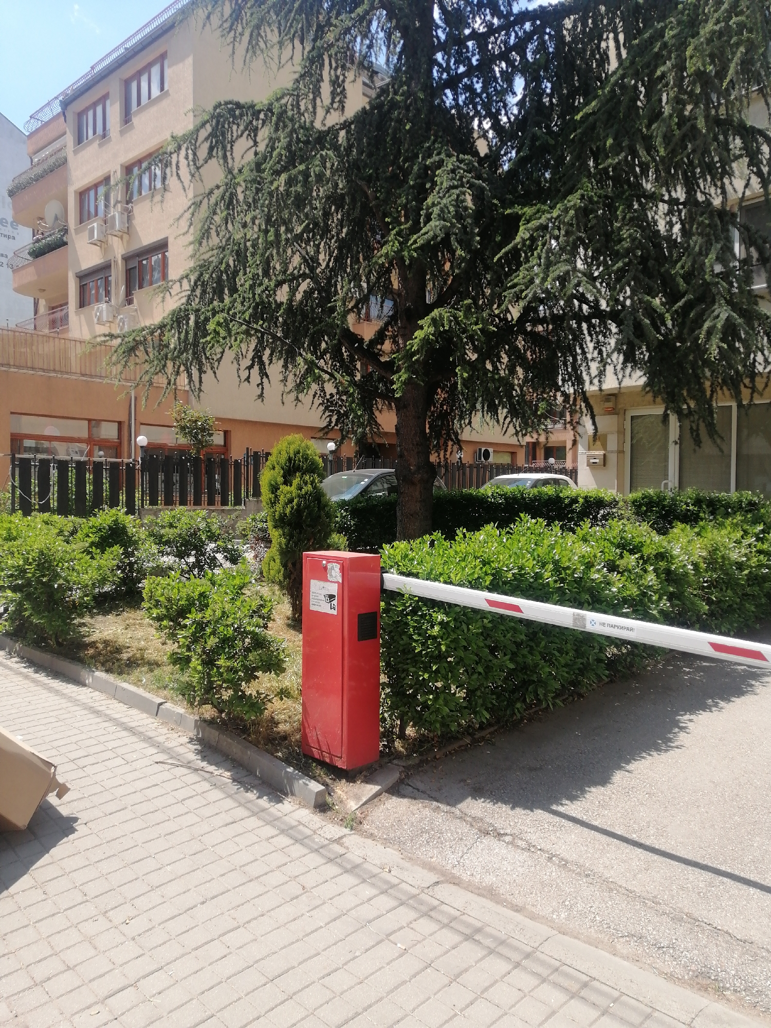 The image shows a street scene, possibly in Sofia, Bulgaria. A red metal box with a white and red striped barrier arm stands at the edge of a hedge. A cardboard box is visible in the lower left corner. Behind the barrier, there is a landscaped area with bushes, a small tree, and a fence. Two cars are parked in the background. A multi-story building with a light tan facade, multiple windows, and some air conditioning units is visible in the background. The street is paved with interlocking concrete pavers. The scene is well-lit, suggesting it is daytime.