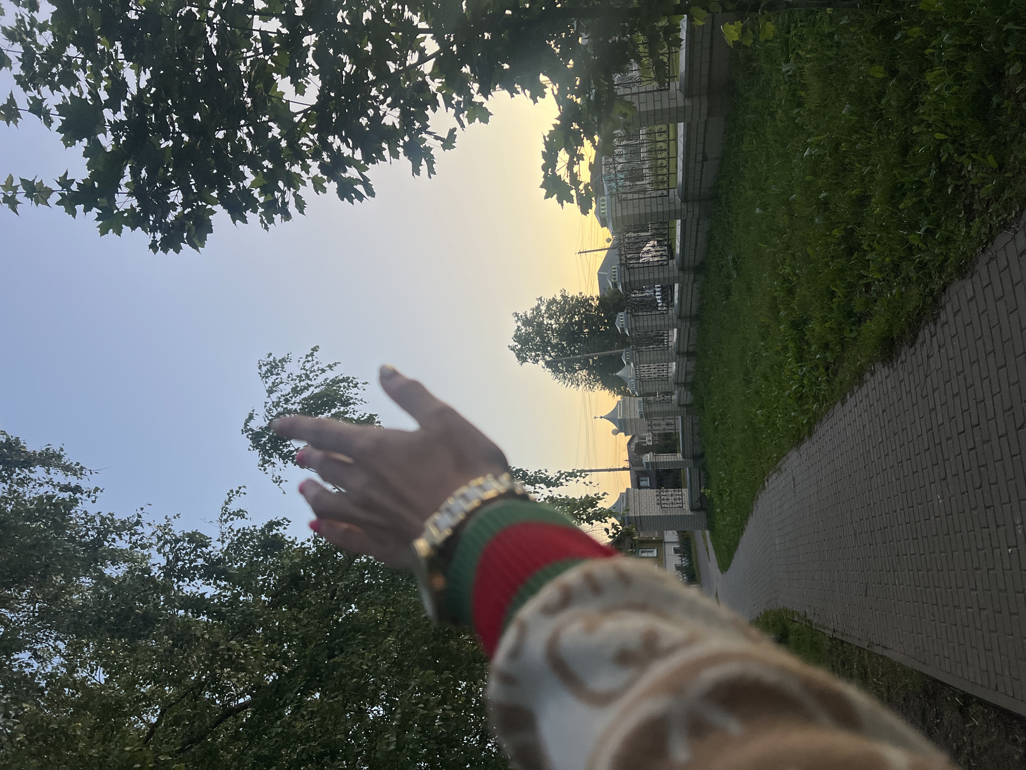 A first-person perspective shot shows a person's arm and hand reaching towards the sky. The arm is adorned with a bracelet and a sleeve featuring a distinctive green and red stripe pattern, along with a beige fabric with an embossed pattern. The hand, with a touch of pink nail polish, is extended upwards, as if reaching for something in the sky or gesturing.
The scene is set outdoors, with lush green trees framing the upper left of the image. In the background, a paved walkway runs alongside a grassy area, bordered by a low fence and a series of buildings that appear to be houses or residential structures with light-colored walls and dark roofs. The sky is a pale blue, transitioning to a soft yellow or orange hue towards the horizon, suggesting it's either early morning or late evening. The lighting implies a clear, calm day.
The image evokes a sense of freedom, aspiration, or connection with nature. The focus on the hand reaching upwards, combined with the expansive sky and the natural elements, creates a feeling of hope or wonder. The background details of the walkway and buildings suggest an urban or suburban setting within Krasnoe-na-Volge, Russia, possibly a park or a residential street near greenery. No visible text is present in the image.