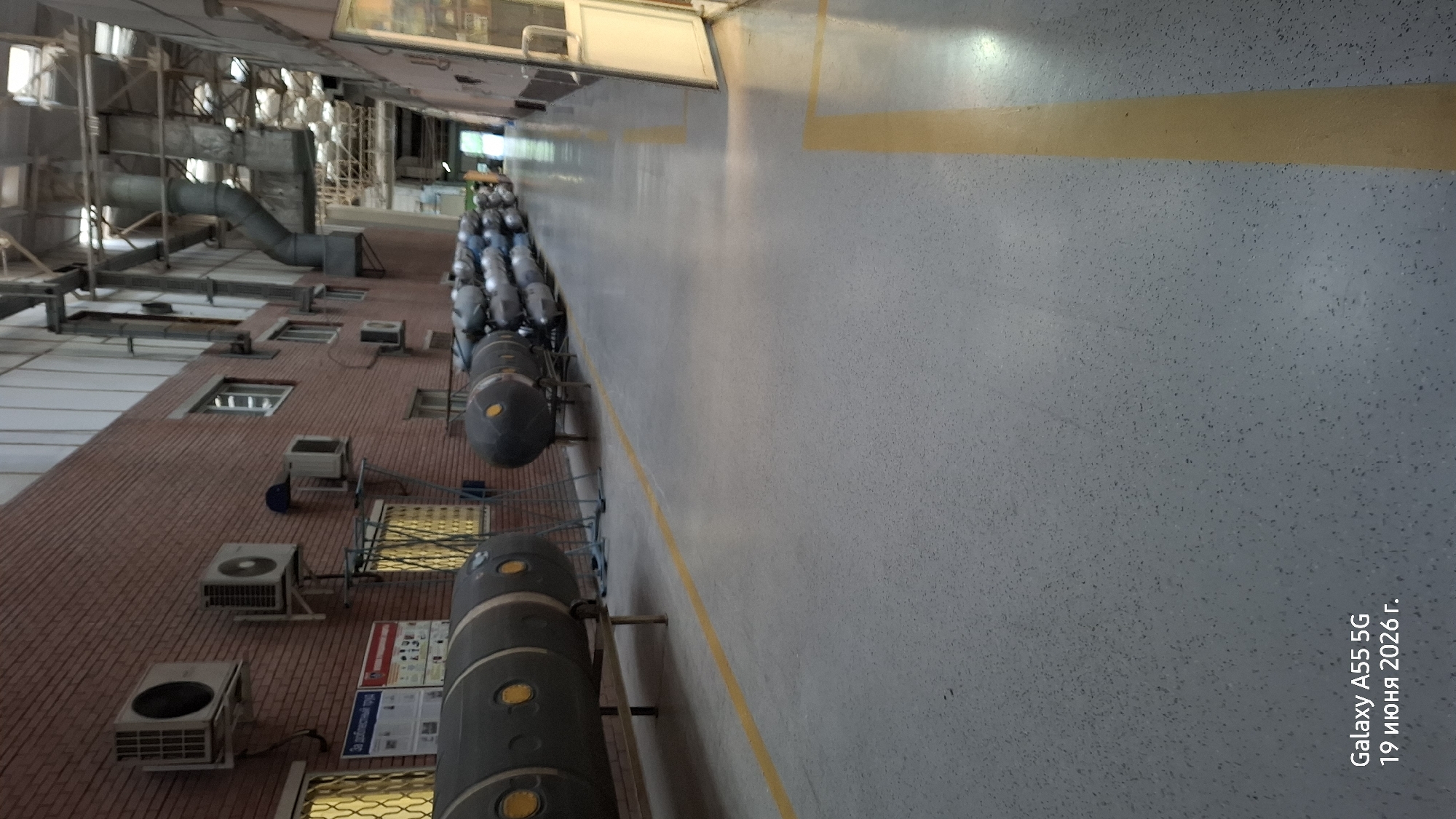 The image shows the interior of a long, industrial building, possibly a factory or warehouse. The floor is a light gray speckled material with yellow lines marking pathways. On the left, a brick wall runs along the length of the building, with several air conditioning units mounted on it. Below the wall are windows with yellow grates and some posters. In front of the wall, large, cylindrical objects are stored on metal racks. These objects are dark gray and appear to be some kind of tanks or canisters, some with yellow circular markings. Further down the building, more of these cylindrical objects are lined up, extending into the distance. The ceiling is high and features exposed pipes and ductwork. On the right side of the image, there is a white door and part of a glass partition. The overall impression is one of an active industrial space with items in storage. The lower left corner of the image contains text indicating "Galaxy A55 5G" and a date in Russian: "19 июня 2026 г.".