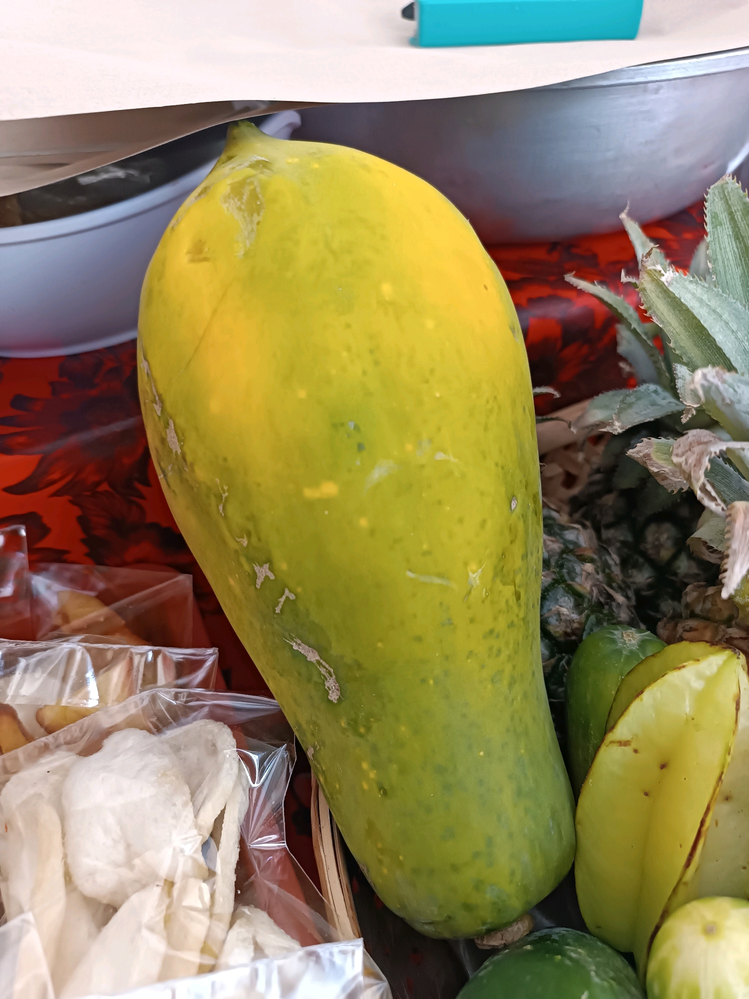 A still life photograph captures a close-up of a collection of fruits and snacks. Dominating the center is a large, elongated papaya, its skin a gradient of yellow and green with speckled imperfections. To its right, the spiky crown of a pineapple is visible, along with slices of starfruit, revealing their distinct five-pointed shape. In the foreground, plastic-wrapped packets of white, puffed snacks, possibly krupuk, are neatly arranged. The background is a blur of red floral patterns and a hint of a silver bowl. The scene appears to be set outdoors, likely at a market stall in Tabanan, Indonesia, given the tropical fruits and common snacks. The lighting is natural, casting soft shadows. There are no people or animals present in the frame.