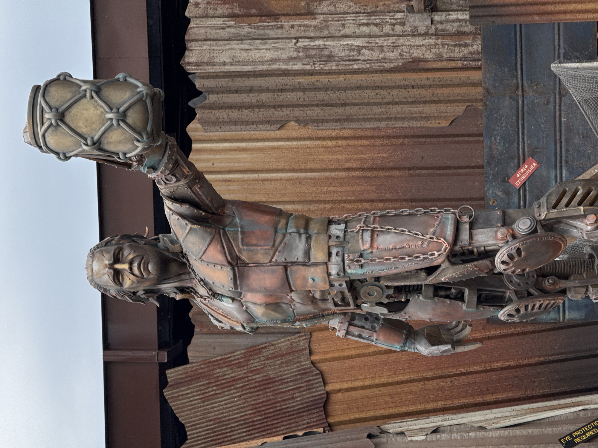 A steampunk-style sculpture of a man holding a drum is affixed to a corrugated metal wall. The sculpture is composed of various metal pieces, chains, and gears, giving it a mechanical appearance. A small red label with white lettering, partially obscured, can be seen on the wall next to the sculpture. A sign with the words "EYE PROTECTION REQUIRED" is barely visible in the bottom right corner. The location is Tongzhou District, China.