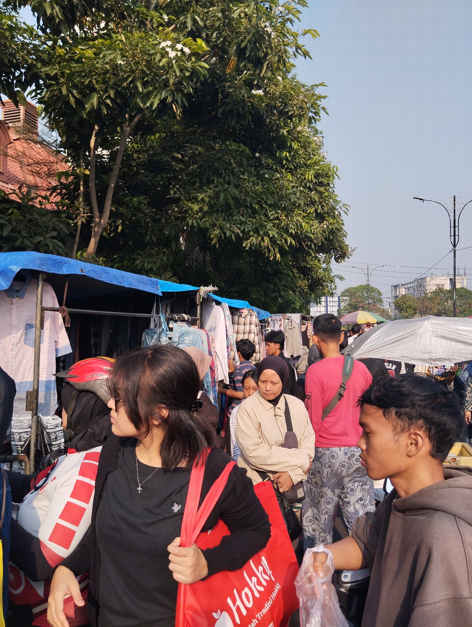 This image depicts a busy outdoor market scene in Surabaya, Indonesia. In the foreground, a young woman wearing sunglasses and a black t-shirt is holding a red bag. Beside her, a young man in a grey hoodie is holding a plastic bag. Behind them, several other individuals are visible, including a woman in a beige jacket and headscarf, and a man in a pink long-sleeved shirt. The market features stalls with various clothing items displayed under blue tarpaulins, and other stalls with silver and colorful umbrellas. Trees and a street light are visible in the background, indicating an urban setting. The sky is clear and blue, suggesting daylight.
